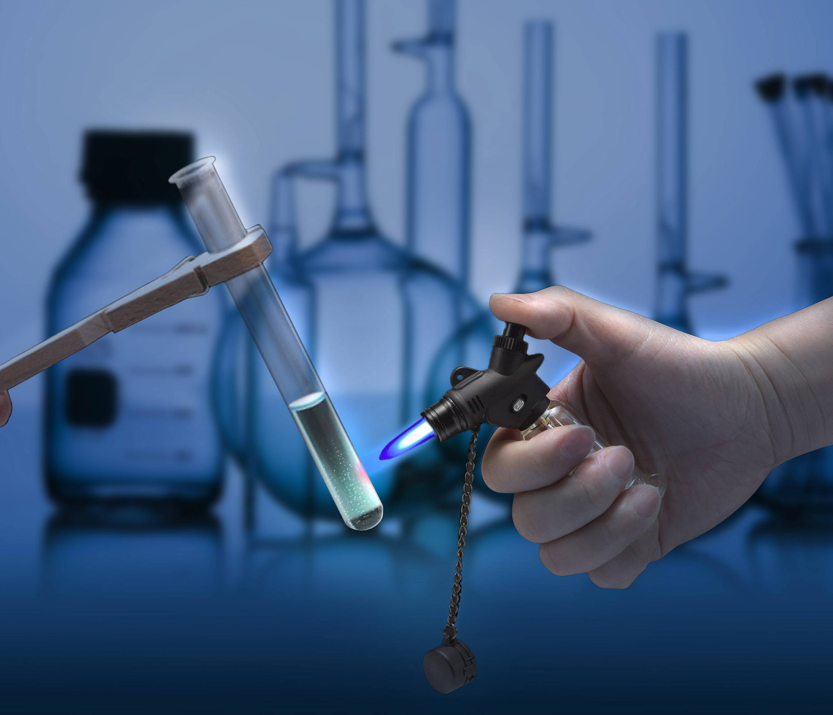 Dłoń trzyma palnik, który podgrzewa probówkę z cieczą. W tle niewyraźne są butelki laboratoryjne i probówki.