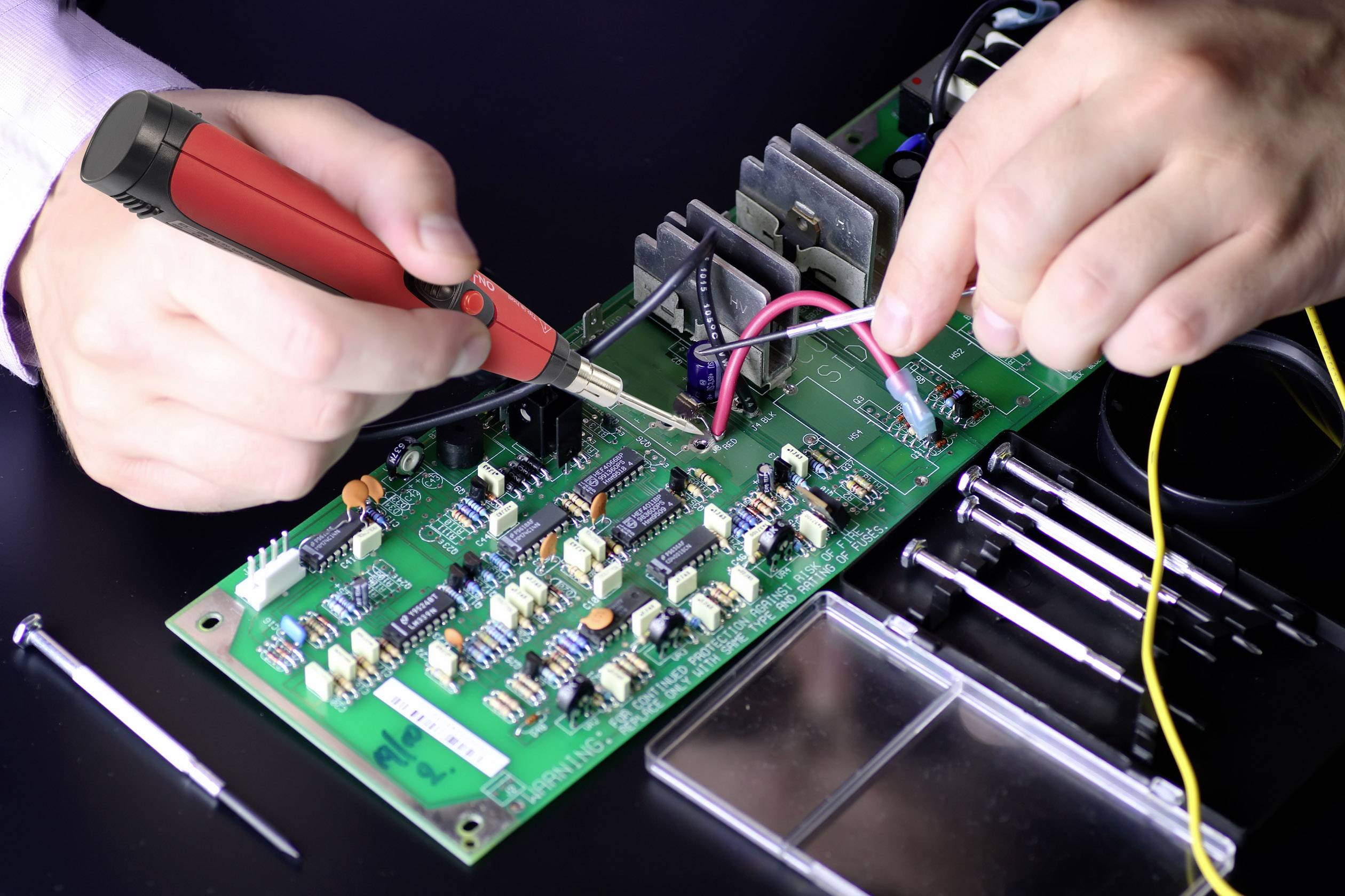 Osoba lutuje lutownicą na zielonej płytce drukowanej, otoczona narzędziami.