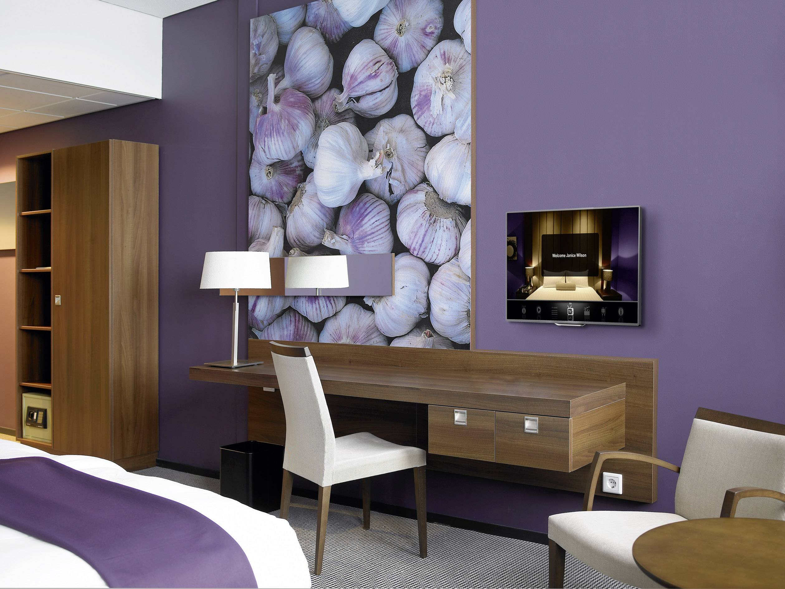 Pokój hotelowy z fioletową ścianą, nad którą wisi obraz z czosnkiem. Drewniane meble, biurko z lampą, telewizor na ścianie, przed nim fotel.