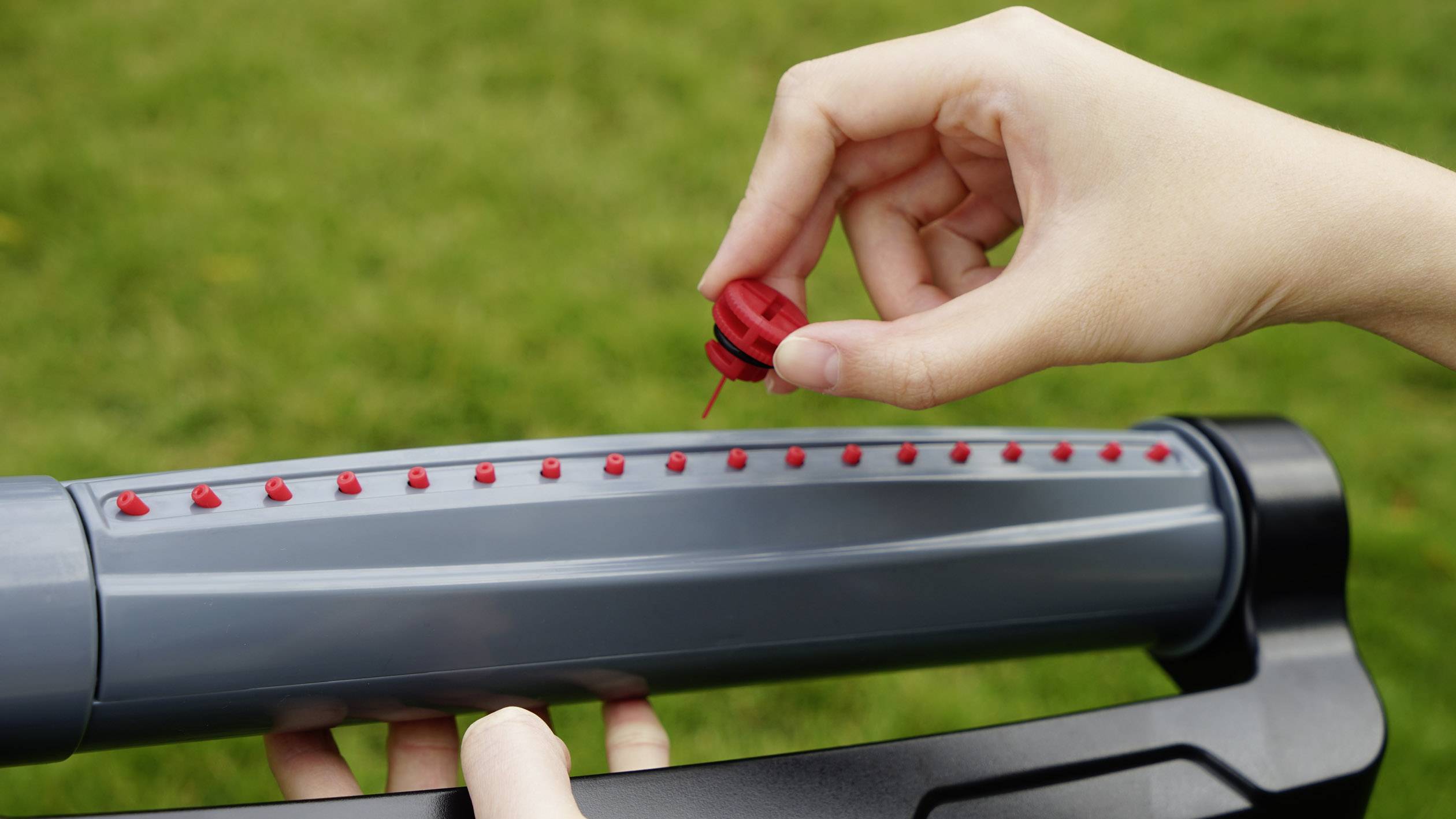 Osoba wkłada czerwony kroplownik do szarego węża ogrodowego do nawadniania. Rozmyty zielony trawnik w tle.