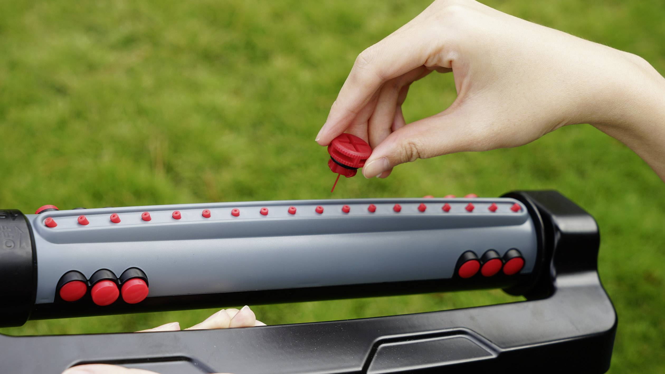 Osoba obraca pokrętło metalowego narzędzia ogrodniczego z czerwonymi przyciskami, które jest ustawiane na zielonej trawie.