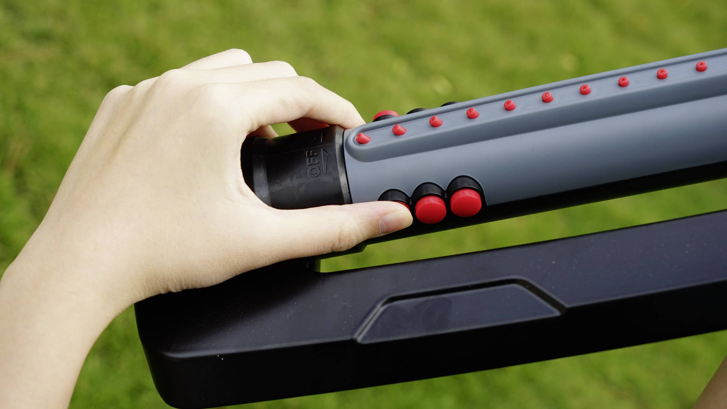 Dłoń trzyma uchwyt składanego chodzika i naciska czerwony przycisk, w tle: trawa.
