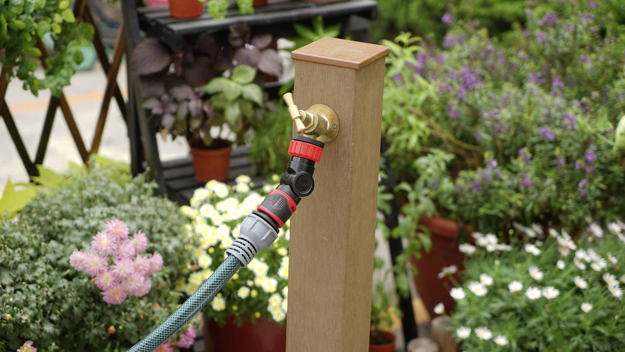 Wąż ogrodowy podłączony do drewnianego słupa, otoczony kwitnącymi roślinami i kwiatami ogrodowymi. Idealny do nawadniania ogrodu.