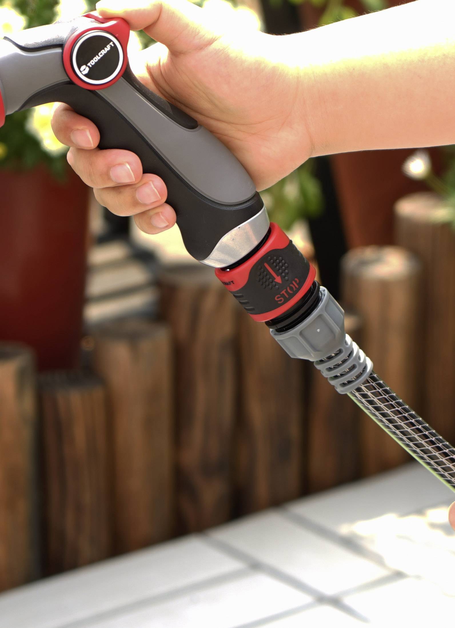 Ręka trzyma dyszę węża ogrodowego z pokrętłem regulacyjnym, w tle widać rozmyte rośliny i drewniane płoty.