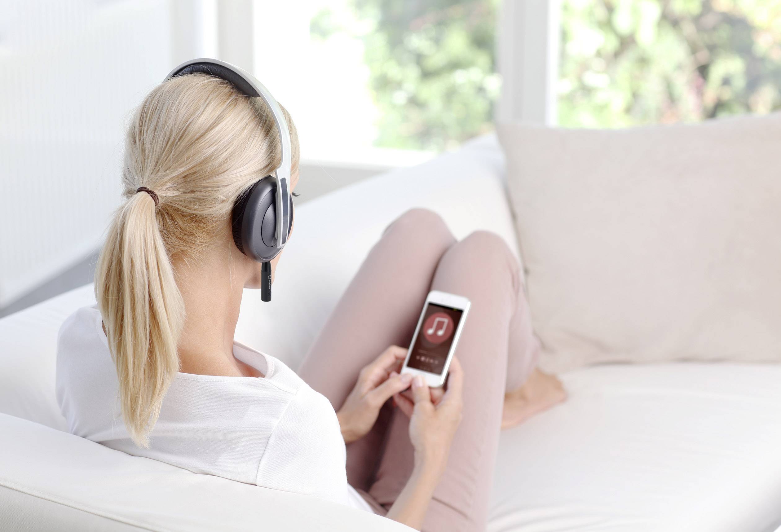 Osoba w słuchawkach siedzi na kanapie i obsługuje smartfona. Na ekranie widoczne jest logo aplikacji muzycznej.