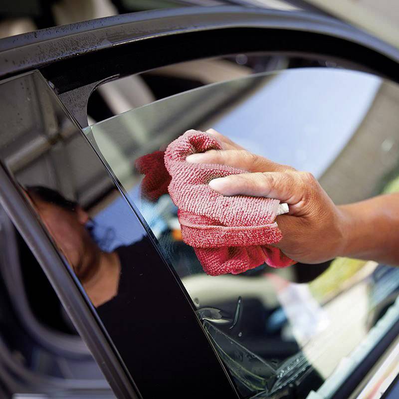 Czerwona ścierka pociera szybę samochodu. Część ramienia osoby jest widoczna, trzymająca szmatkę w świetle słonecznym.