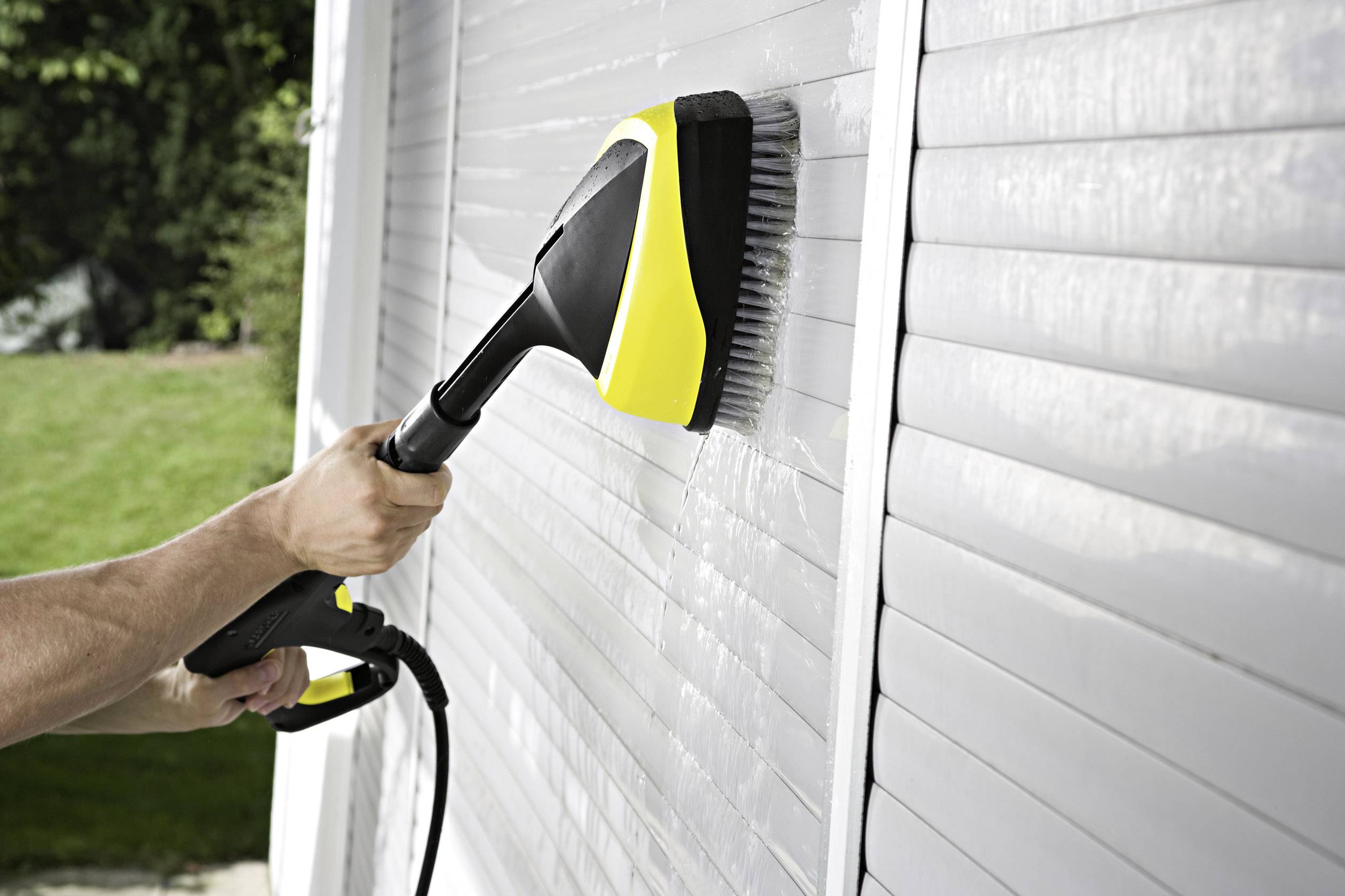 Osoba czyści białą roletę za pomocą myjki ciśnieniowej. W tle widać trawnik i drzewa.