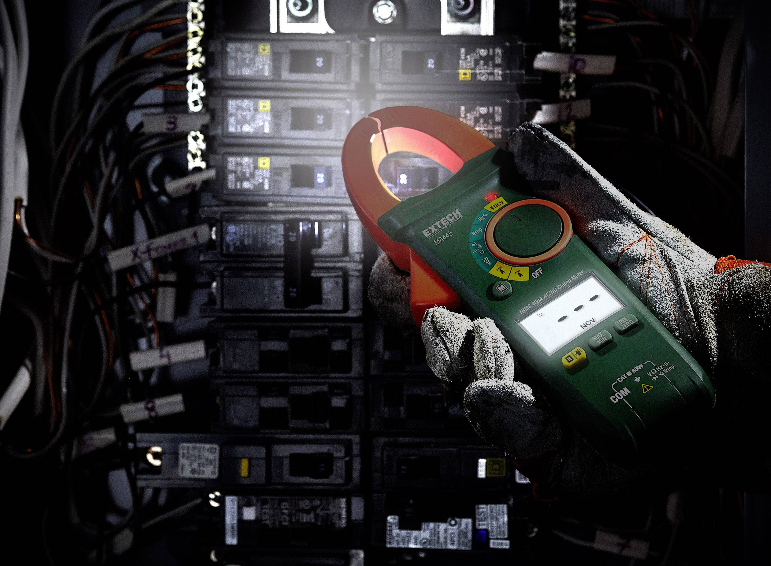 Elektryk trzyma miernik prądu przed otwartą szafką elektryczną z wieloma bezpiecznikami i kablami.