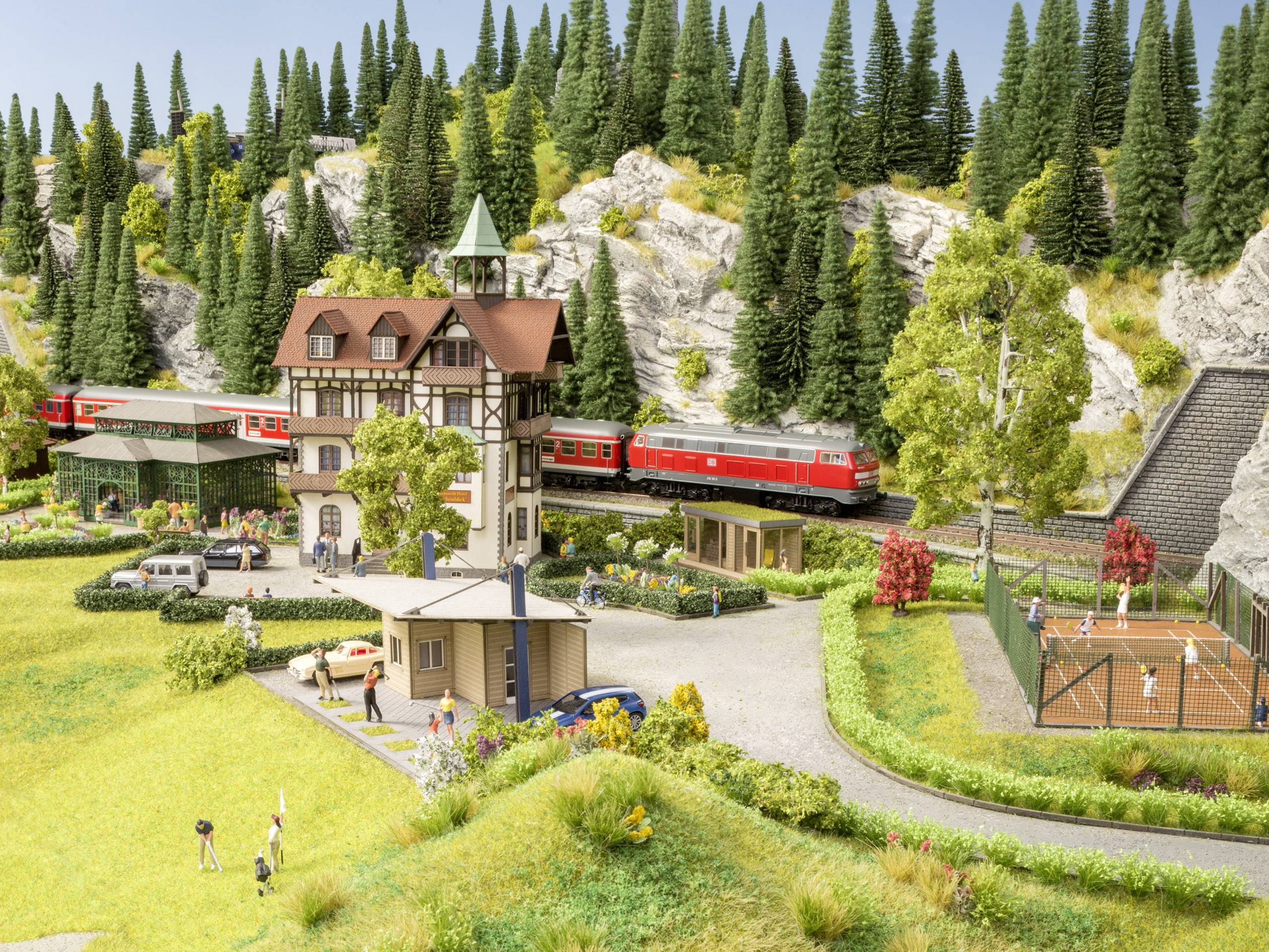 Krajobraz makiety z koleją, górskim tłem i lasem. Czerwony pociąg przejeżdża obok budynku szachulcowego. Na pierwszym planie widać ludzi i kort tenisowy.