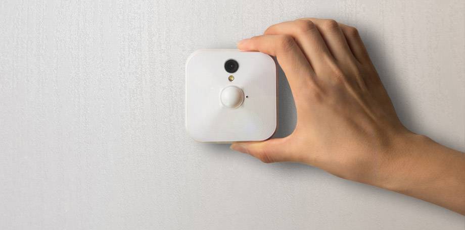 Ręka trzyma mały, kwadratowy czujnik ruchu przy ścianie. Czujnik ma białą powierzchnię i soczewki.