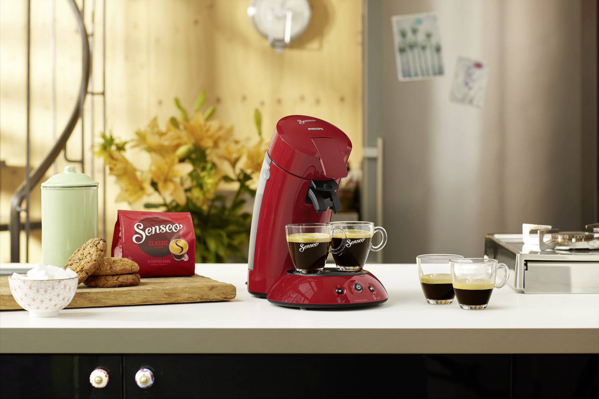 Czerwony ekspres do kawy na blacie kuchennym z dwoma pełnymi filiżankami espresso; w tle kwiaty i opakowanie kawy Senseo.