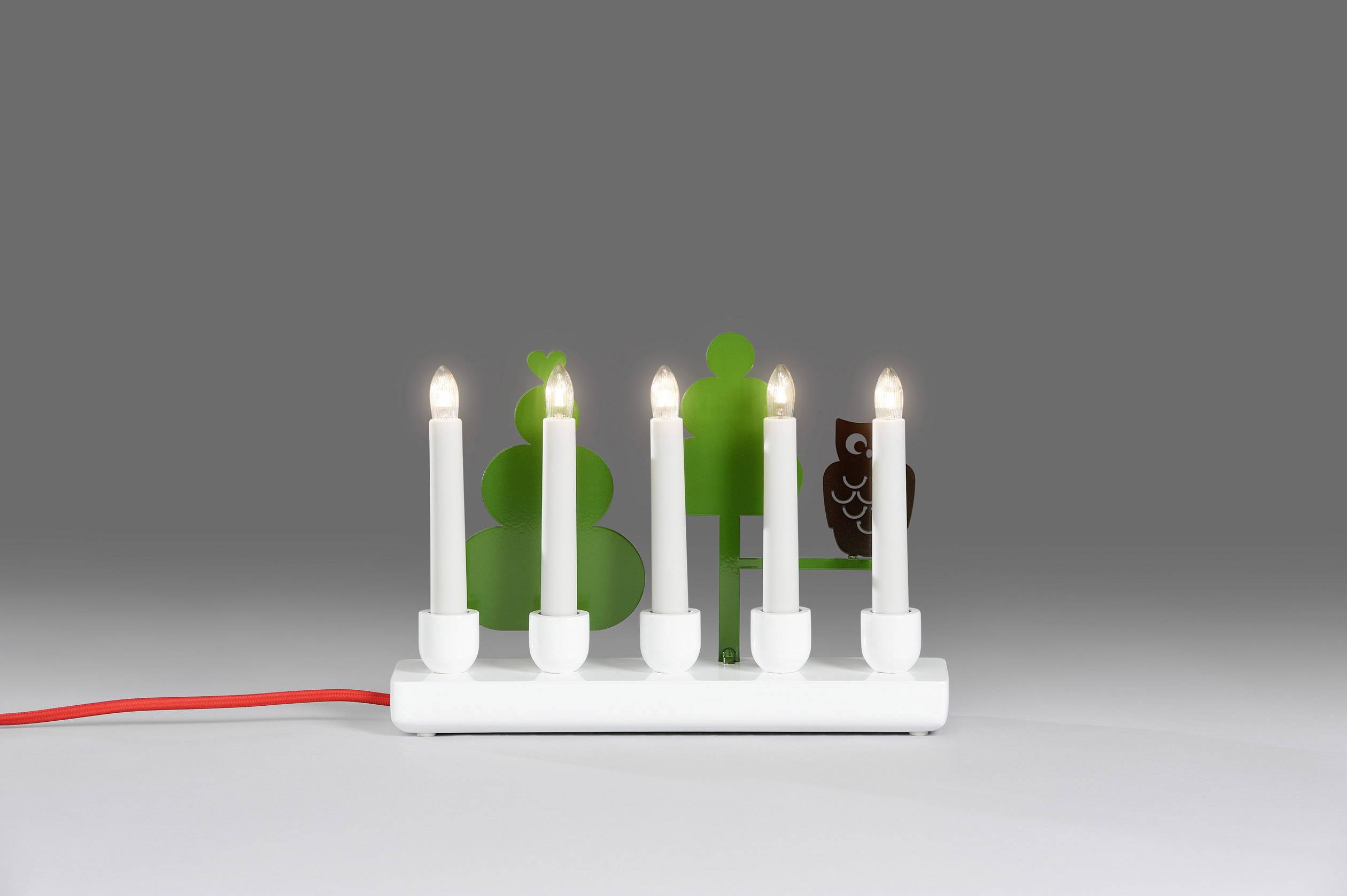 Dekoracyjny świecznik z pięcioma białymi świecami, częściowo otoczony zielonymi i brązowymi figurami drzew, z czerwonym przewodem.