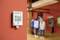 Cyfrowy miernik temperatury i wilgotności na czerwonej ścianie wyświetla '21,5°' i '71,5%'. Na pierwszym planie widać ludzi w pomieszczeniu.