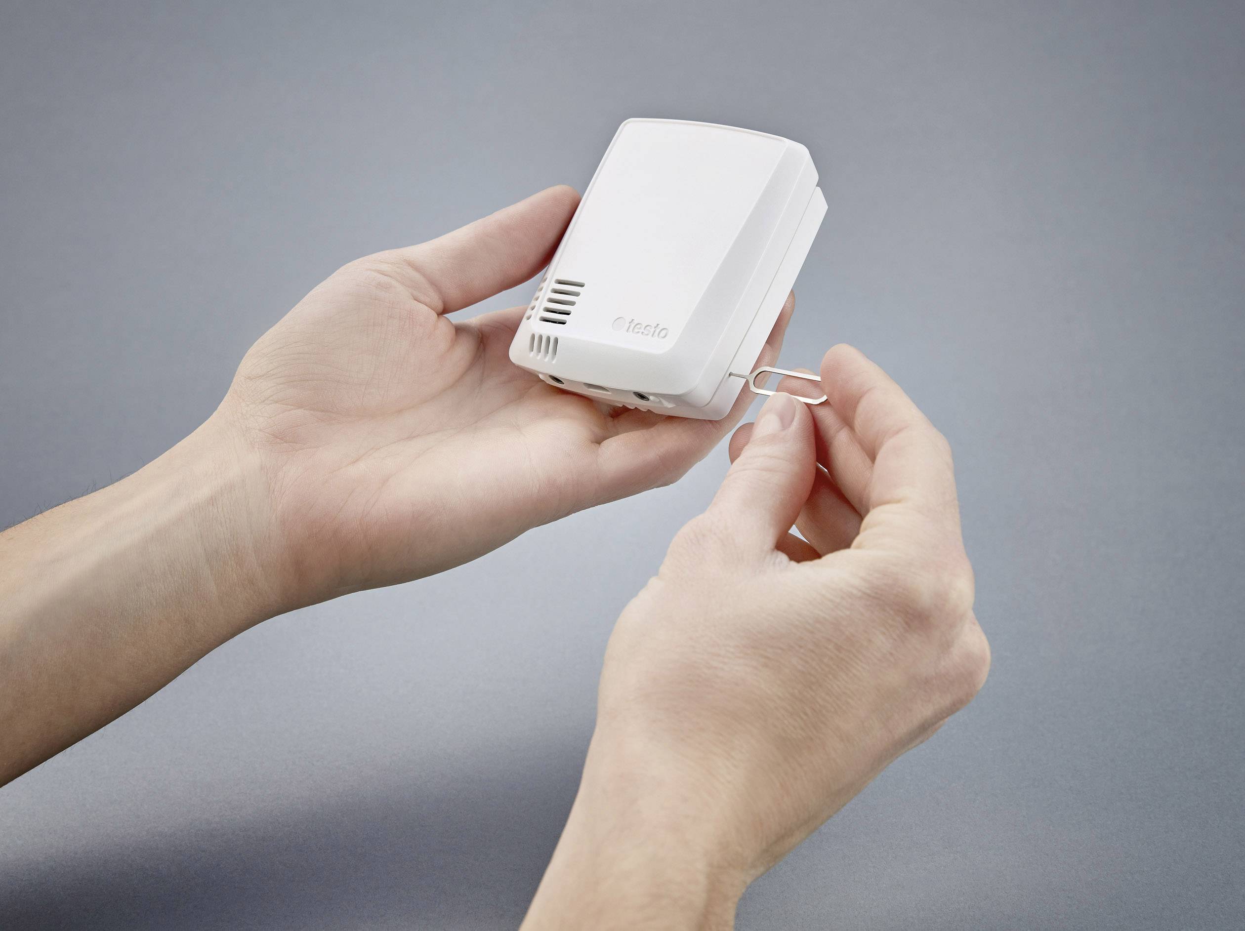 Dwie ręce manipulują małym białym urządzeniem i narzędziem przewodowym. Urządzenie posiada szczeliny wentylacyjne i nazwę marki.