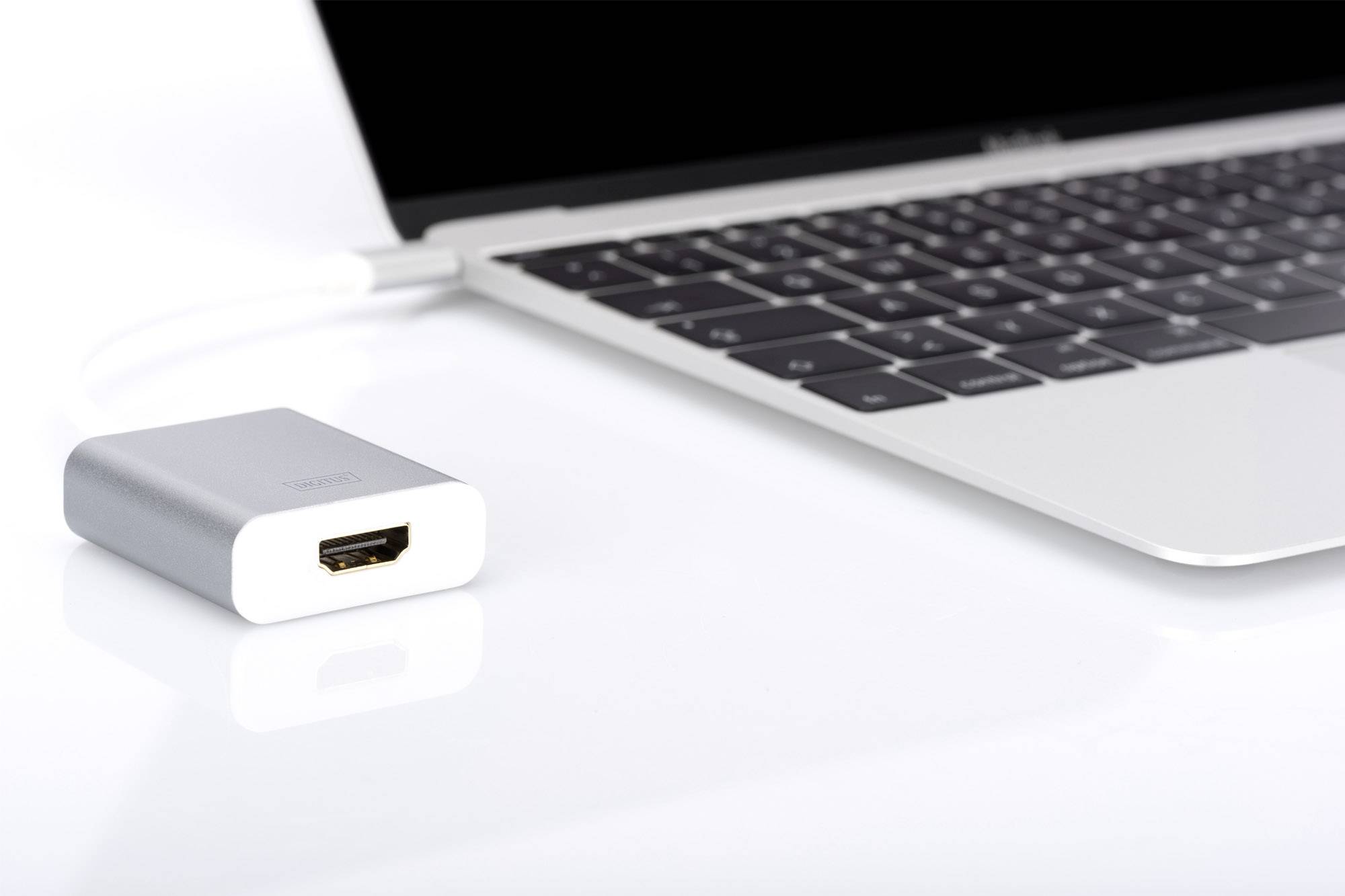 Srebrny adapter HDMI leży obok laptopa z czarną klawiaturą na białym stole. Adapter nie jest podłączony.