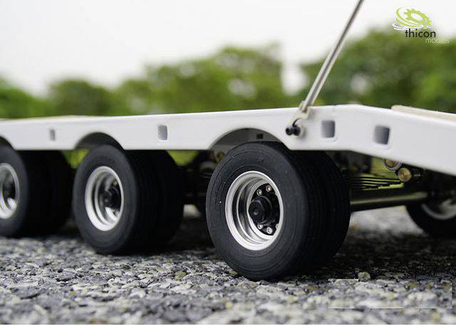 Zbliżenie modelu przyczepy z wieloma kołami na żwirowej powierzchni, w tle rozmyte zielone drzewa.