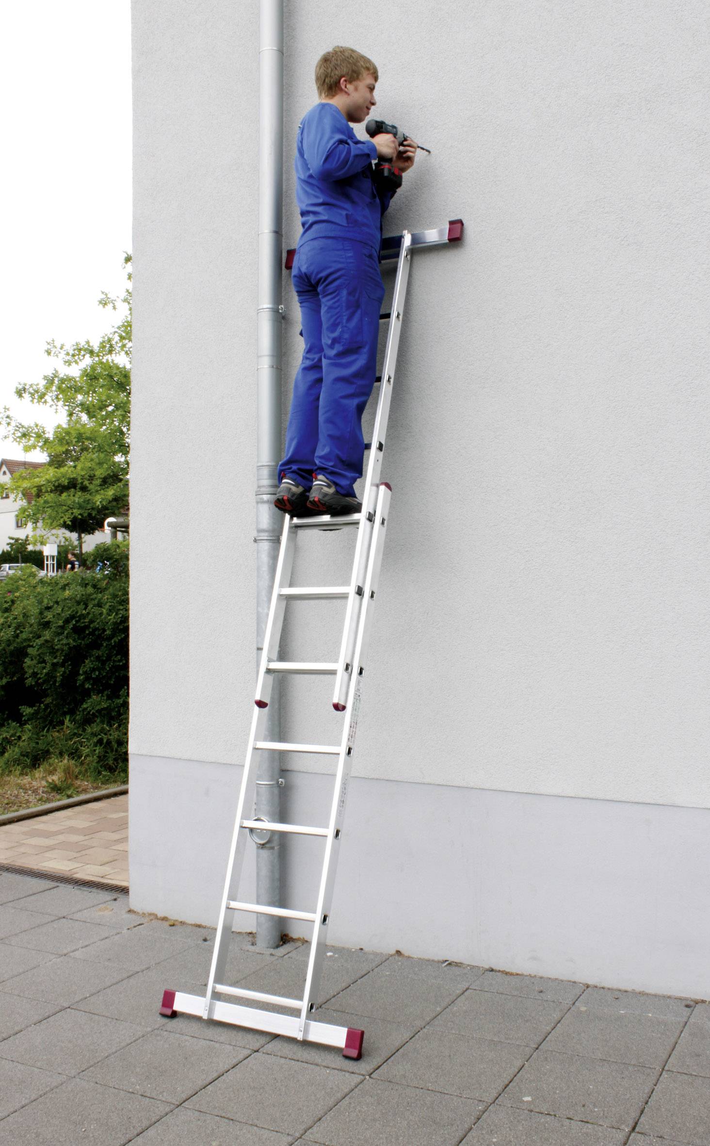 Osoba ubrana w niebieskie ubranie robocze stoi na drabinie obok ściany domu i używa narzędzia, pracując przy ścianie.