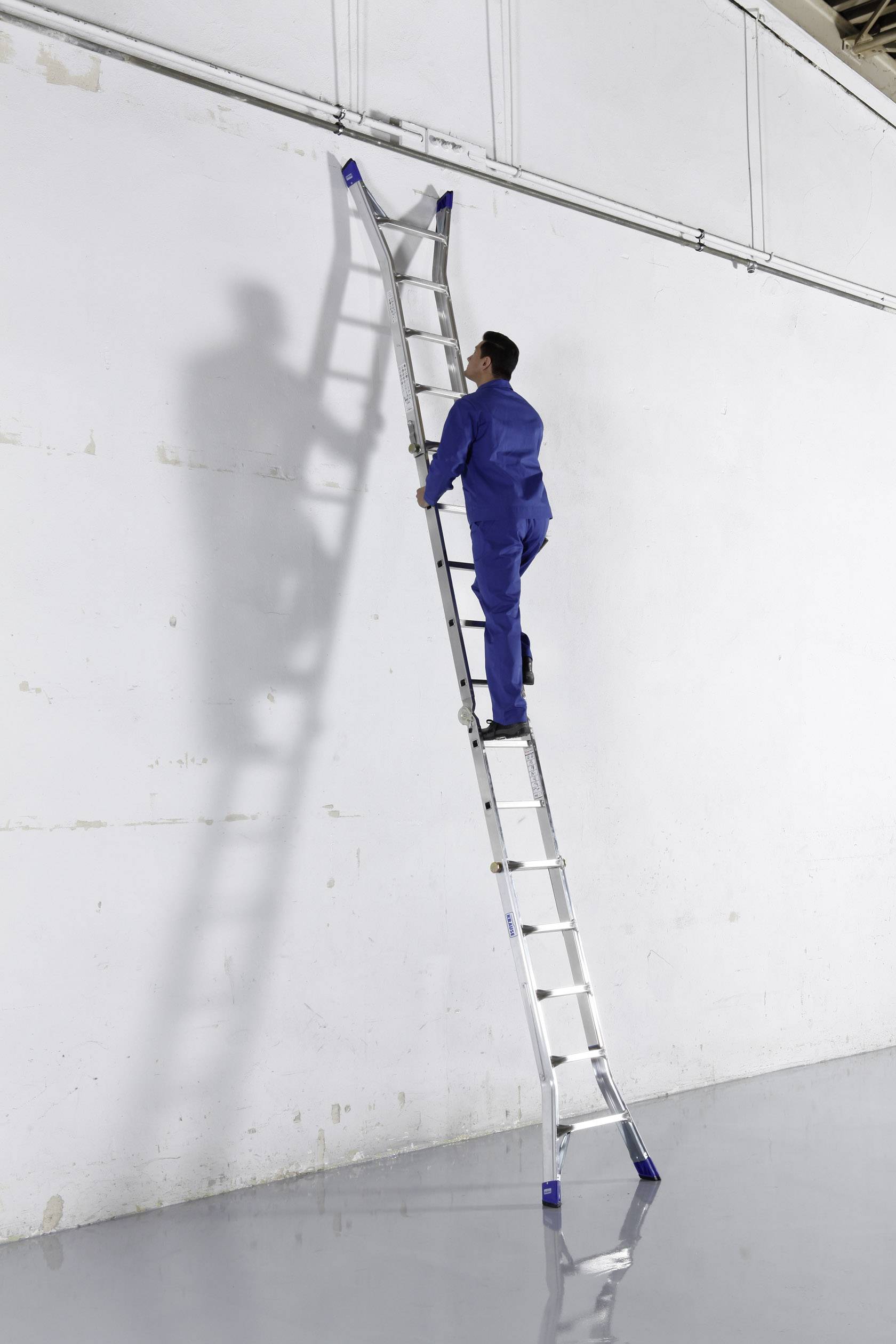 Mężczyzna w niebieskim ubraniu roboczym wspina się po długiej aluminiowej drabinie opartej o białą ścianę w pomieszczeniu przemysłowym.