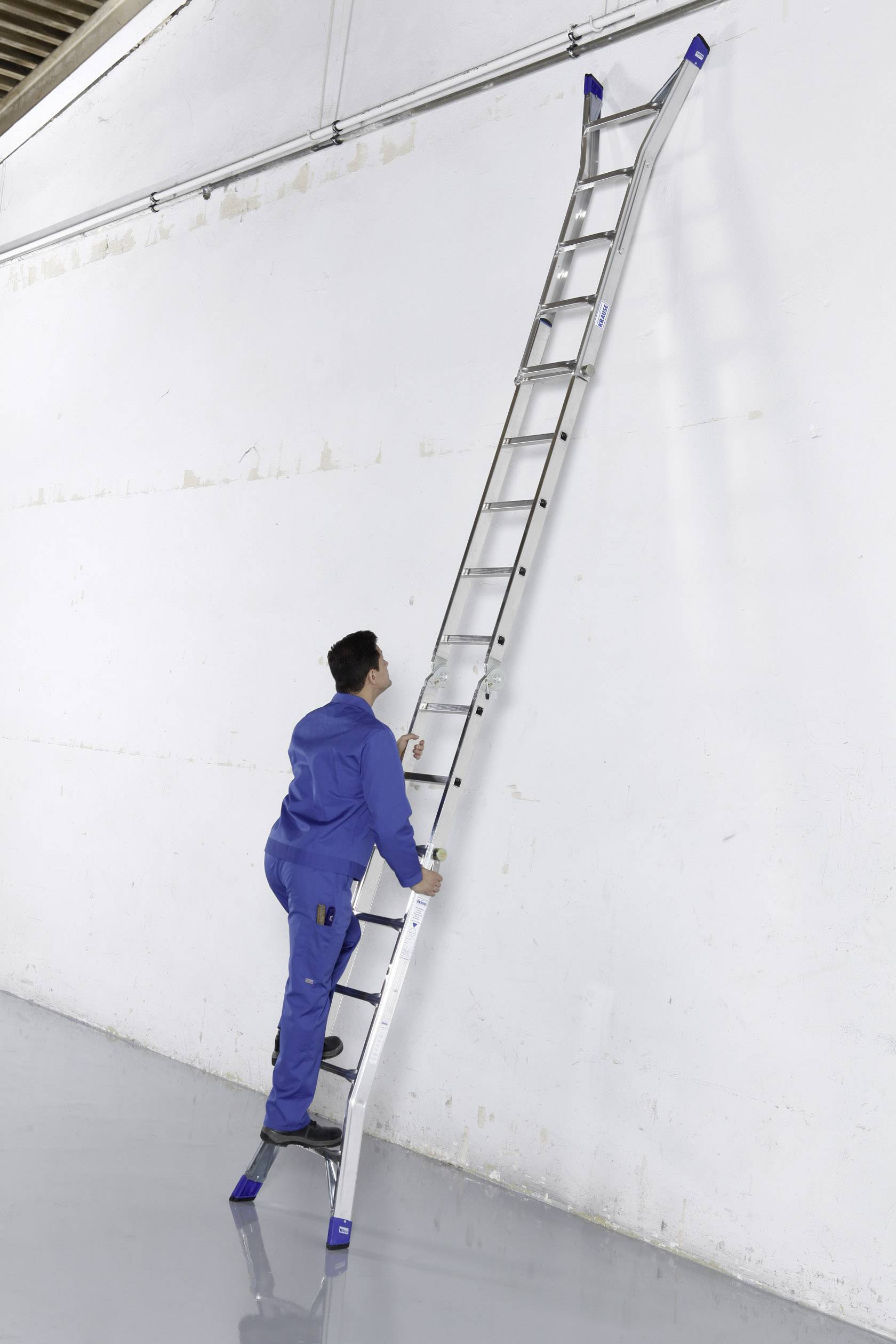 Osoba w niebieskim ubraniu roboczym stoi na długiej aluminiowej drabinie opartej o wysoką białą ścianę.