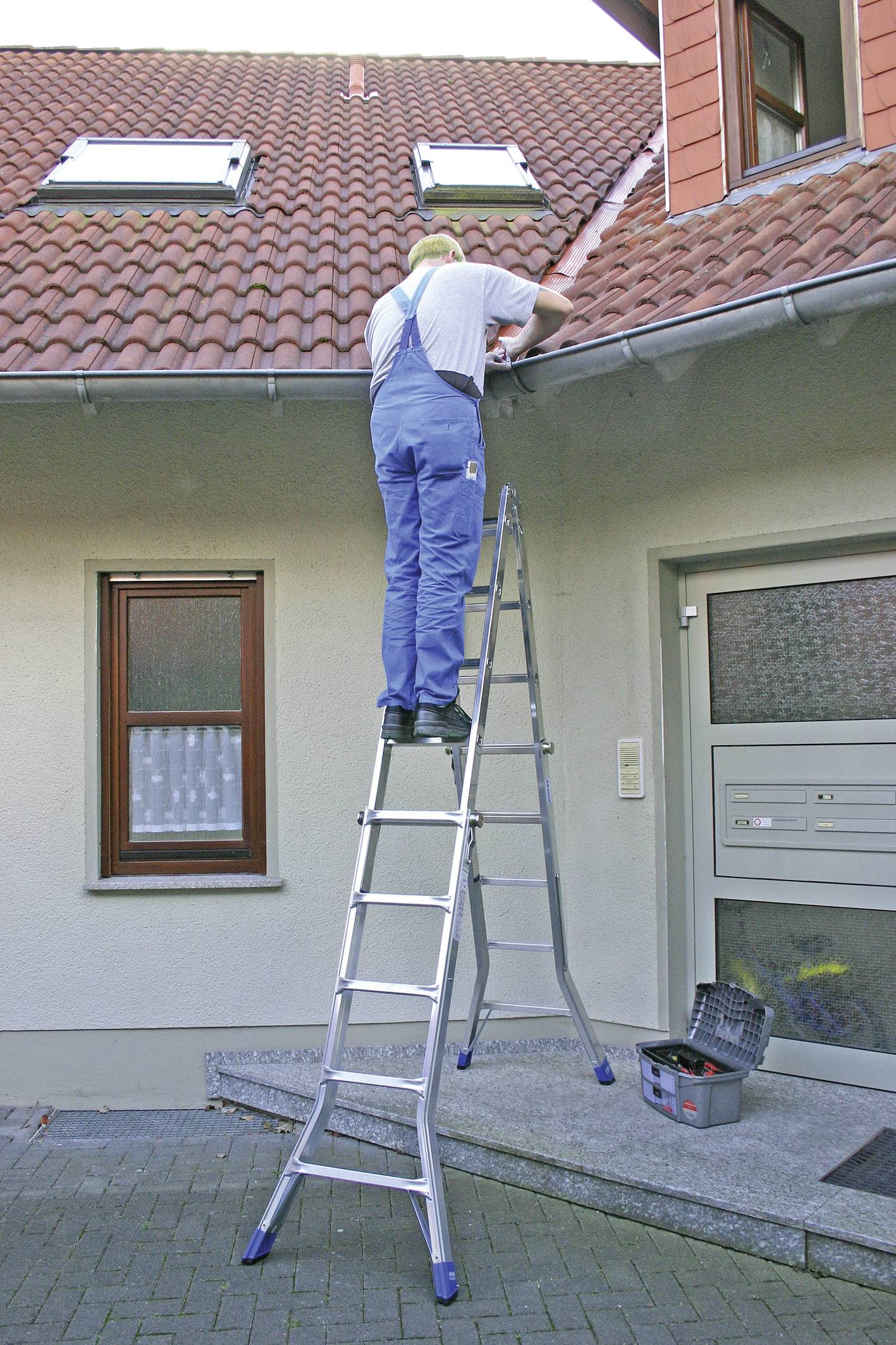 Mężczyzna w niebieskim ubraniu roboczym stoi na drabinie i naprawia dach domu. Na dole stoi skrzynka z narzędziami.