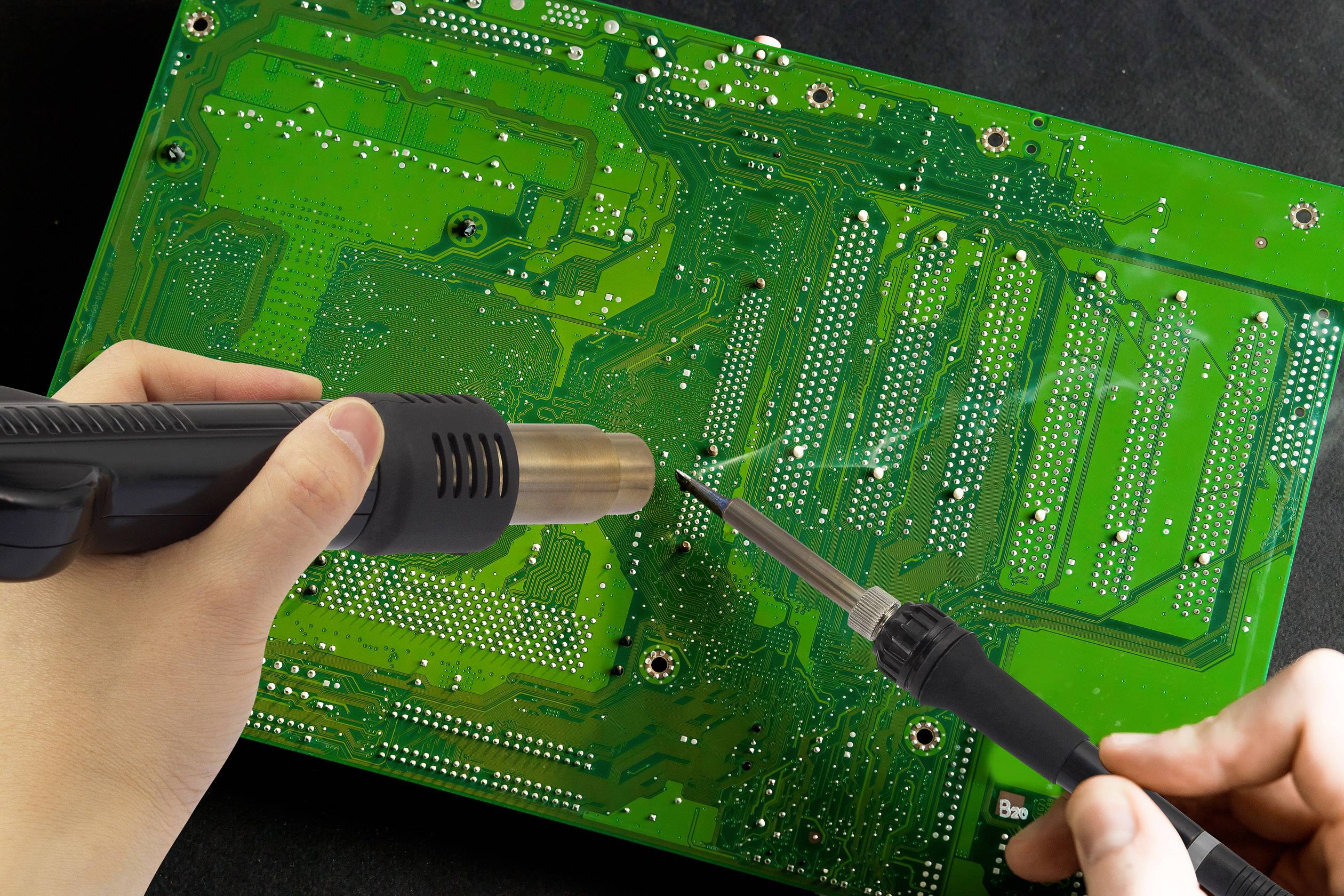 Osoba lutuje zieloną płytkę drukowaną za pomocą lutownicy i gorącego powietrza.