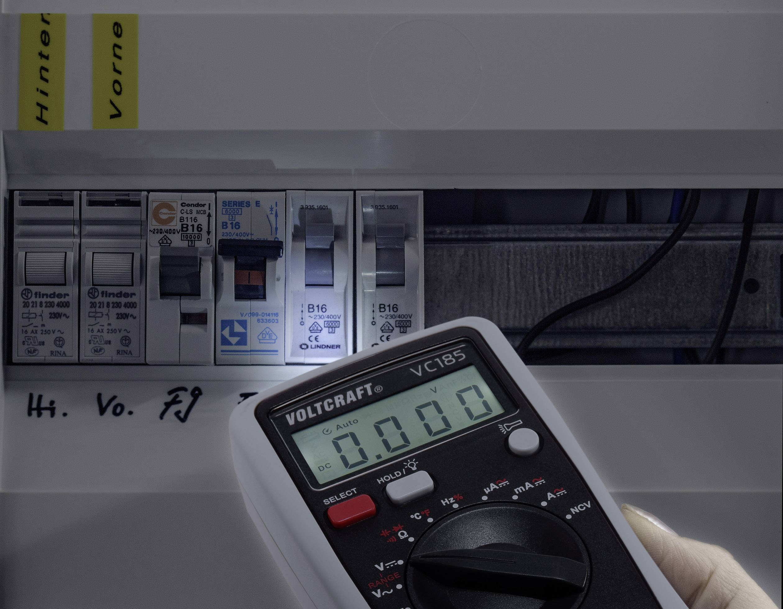 Elektryczna skrzynka bezpieczników z wyświetlaczem wskazującym 0,00 V. Ręka trzyma multimetr.