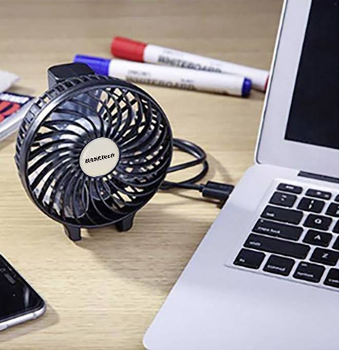 Mały wentylator USB obok otwartego laptopa na biurku. W tle leżą długopisy i smartfon.