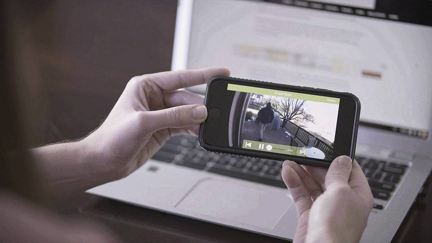 Osoba trzyma smartfon wyświetlający na pierwszym planie obraz z kamery monitoringu na żywo. W tle otwarty laptop.