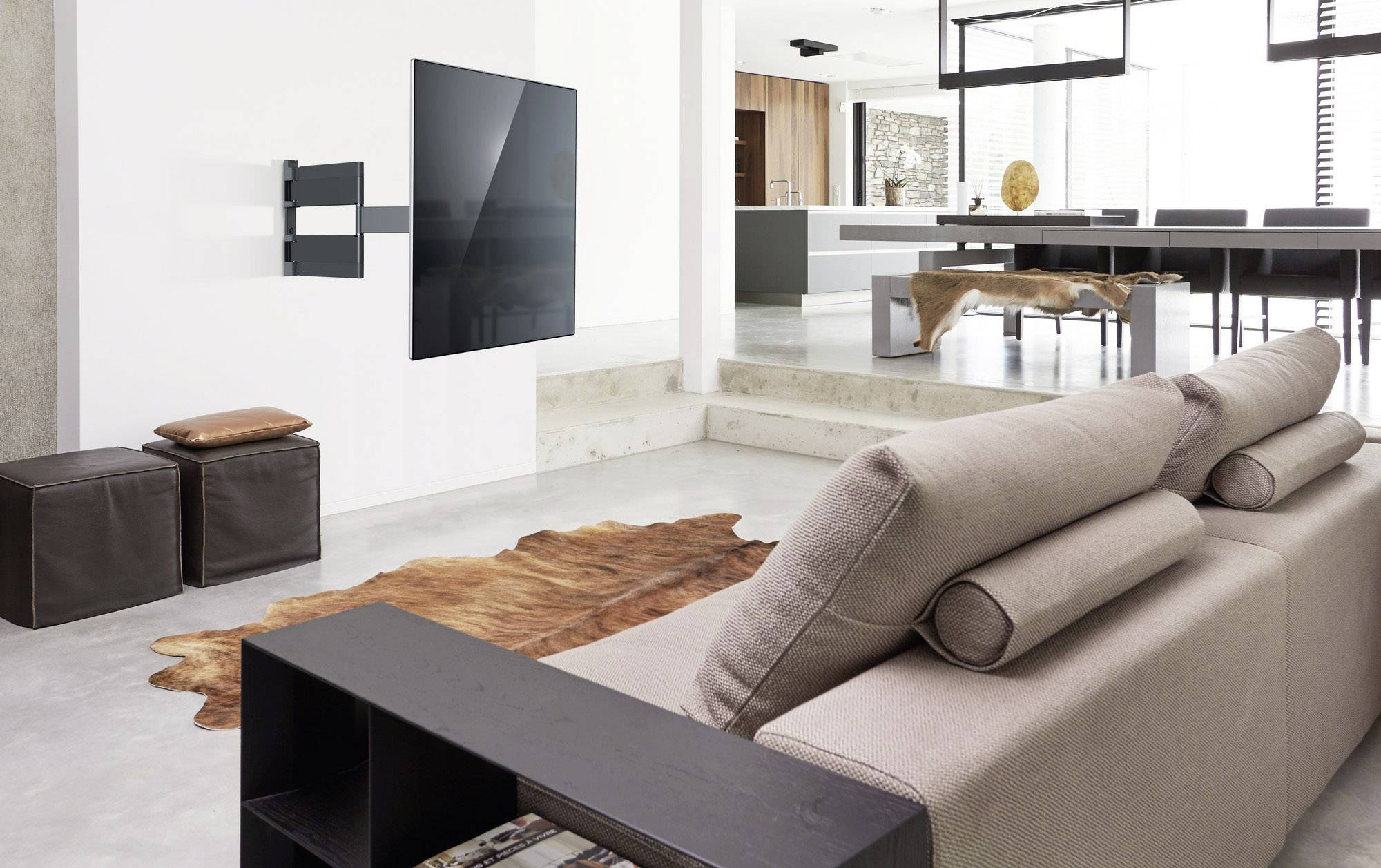 Nowoczesny salon z beżową sofą, telewizorem zamontowanym na ścianie, brązowym dywanem i otwartą kuchnią w tle.