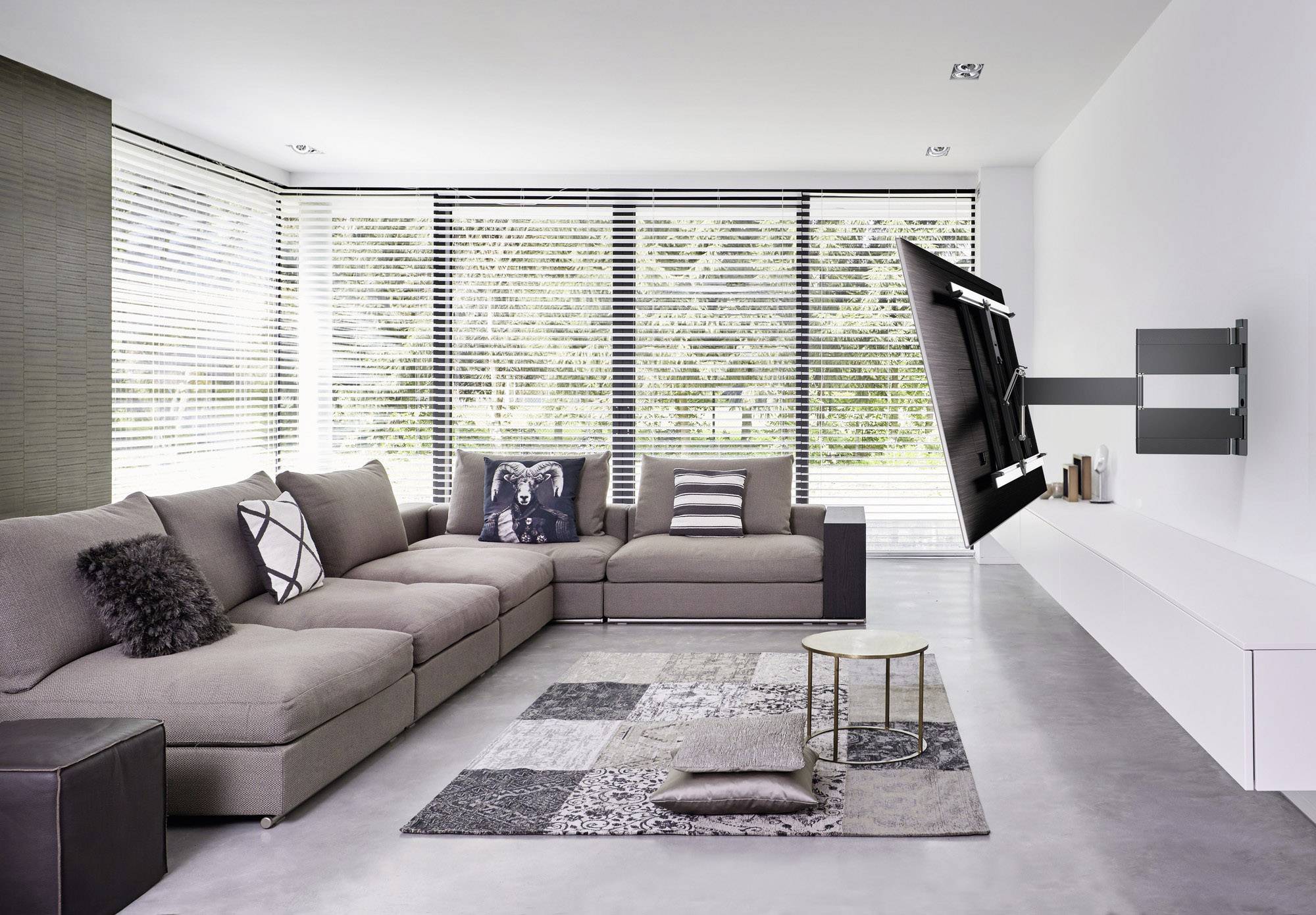 Nowoczesny salon z dużą, szarą sofą, otoczony oknami z żaluzjami. Na ścianie zamontowany jest płaski telewizor.