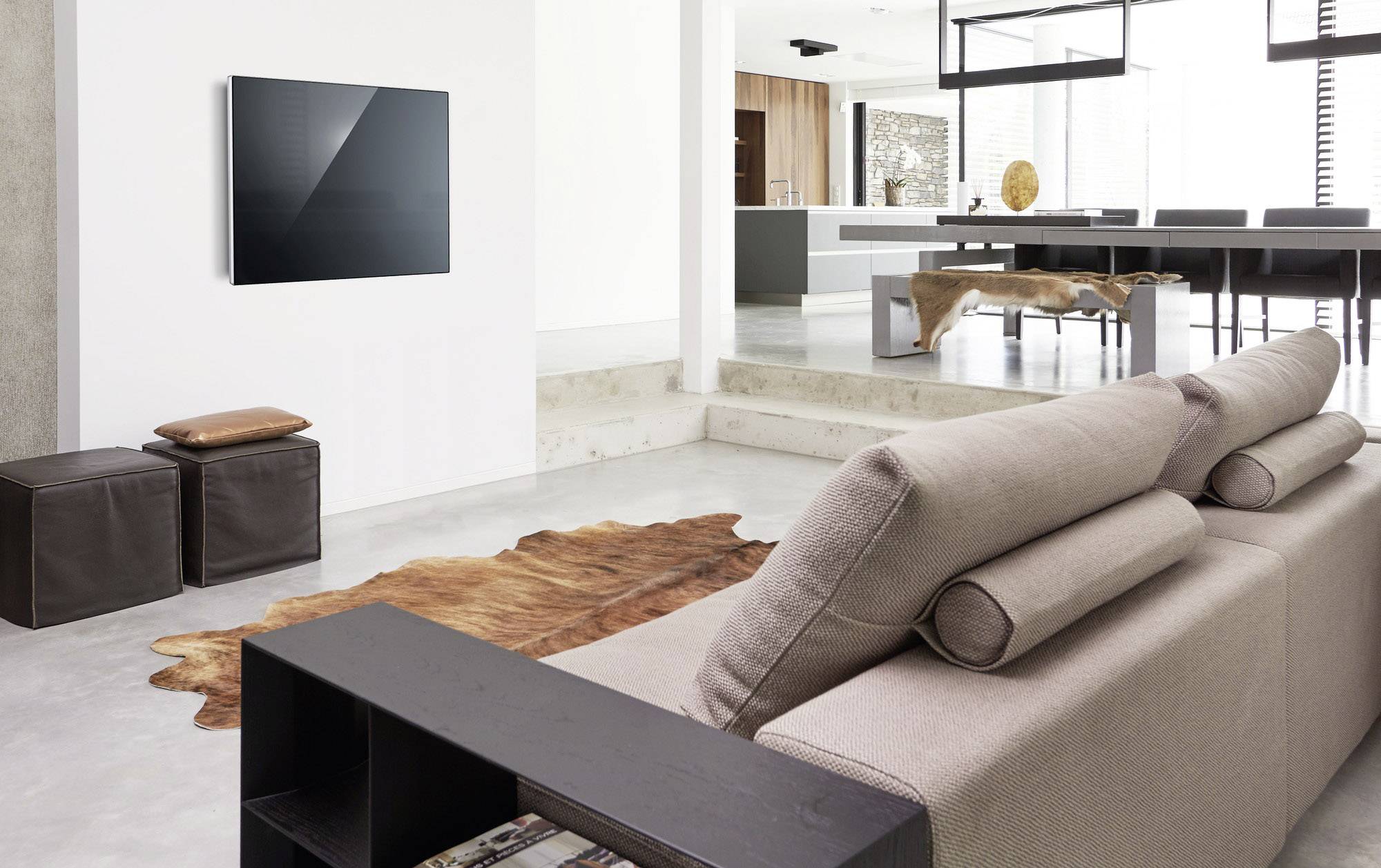 Nowoczesny salon z beżową sofą, drewnianą ławą i telewizorem naściennym. W tle otwarta kuchnia o minimalistycznym wystroju.