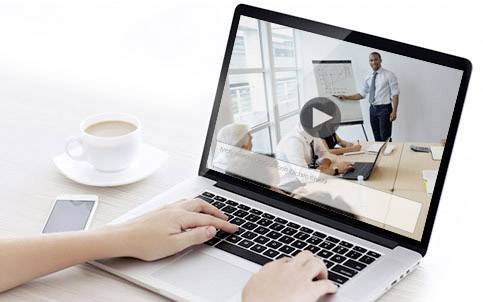 Osoba ogląda wideo edukacyjne na laptopie, które przedstawia prezentację w sali konferencyjnej; obok stoi kubek kawy.