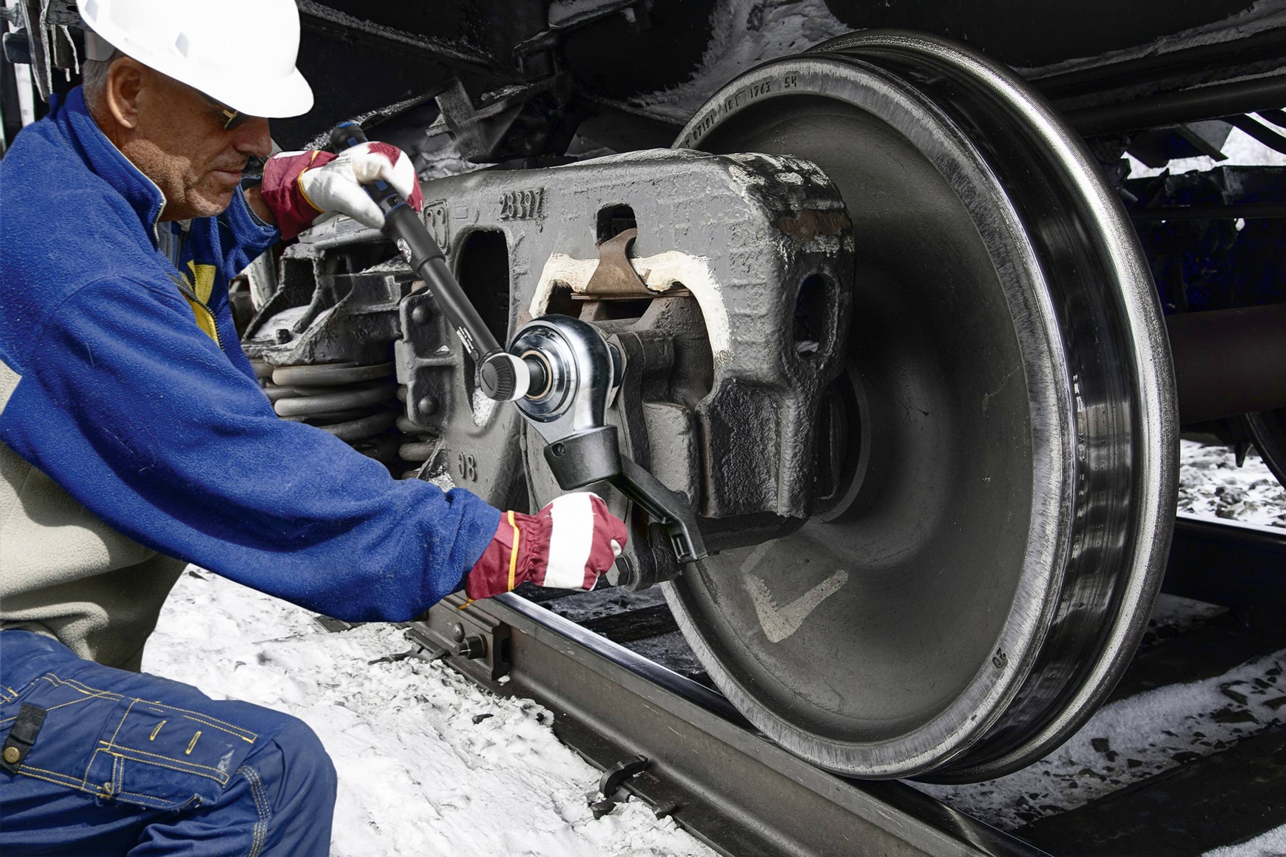 Robotnik w stroju ochronnym naprawia koło wagonu kolejowego narzędziem na wolnym powietrzu.