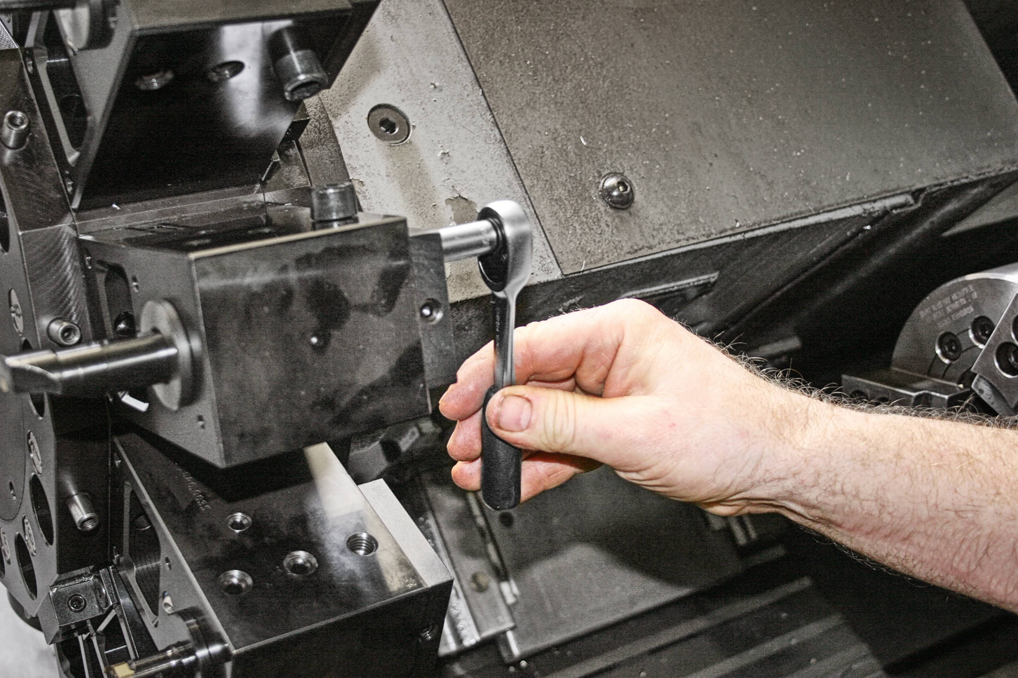 Ręka obsługuje maszynę w środowisku produkcyjnym, prawdopodobnie podczas frezowania lub toczenia elementu metalowego, w pobliżu wrzeciona.