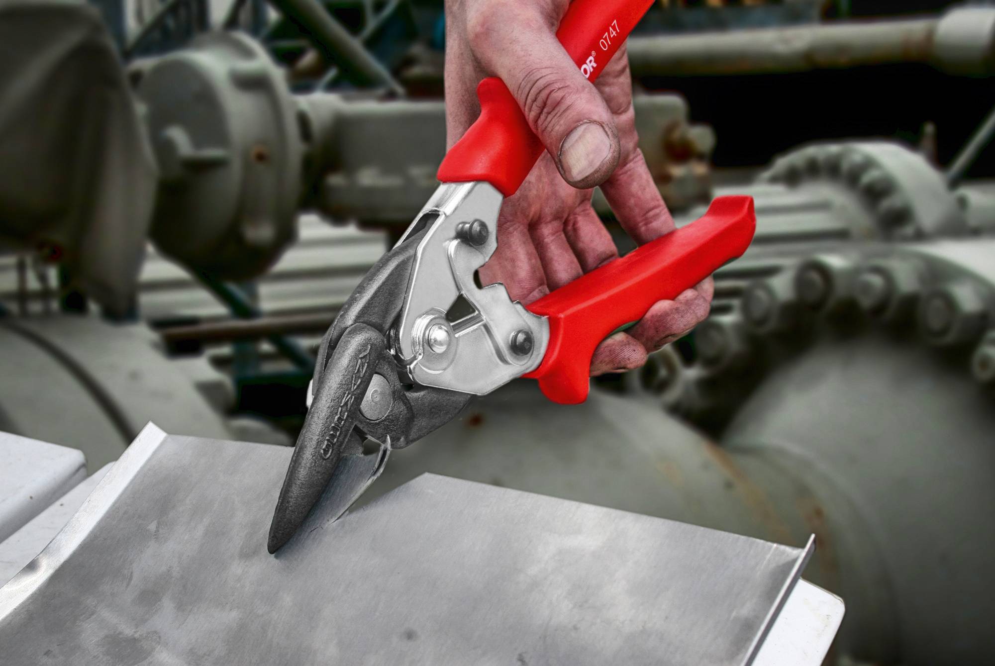 Osoba trzyma czerwone nożyce do blachy i wycina nim kawałek metalu. W tle widoczne są maszyny w przemysłowym otoczeniu.