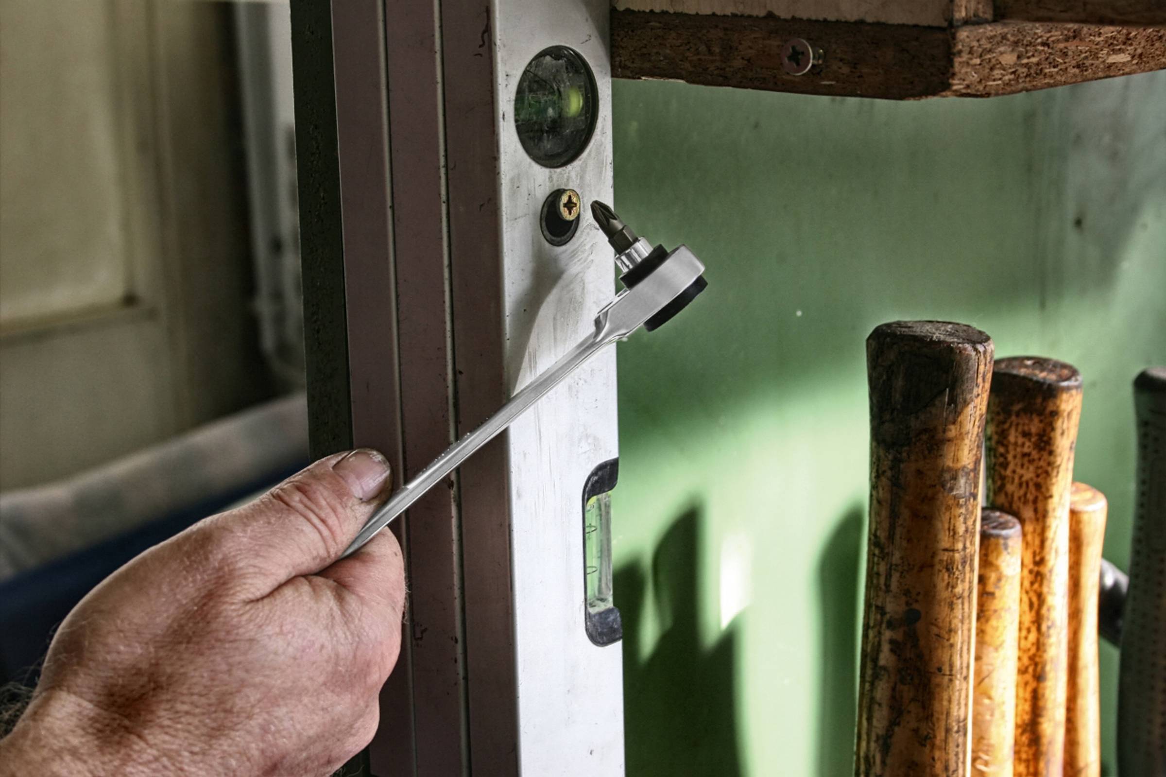 Ręka trzyma narzędzie przy poziomicy, która opiera się o ścianę. W tle stoją drewniane listwy.