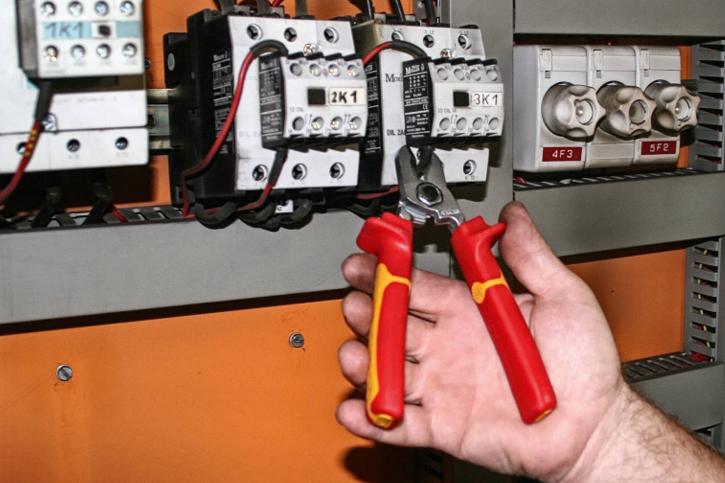 Jedna ręka trzyma szczypce i pracuje przy tablicy rozdzielczej z wieloma elementami elektrycznymi i połączeniami kablowymi.