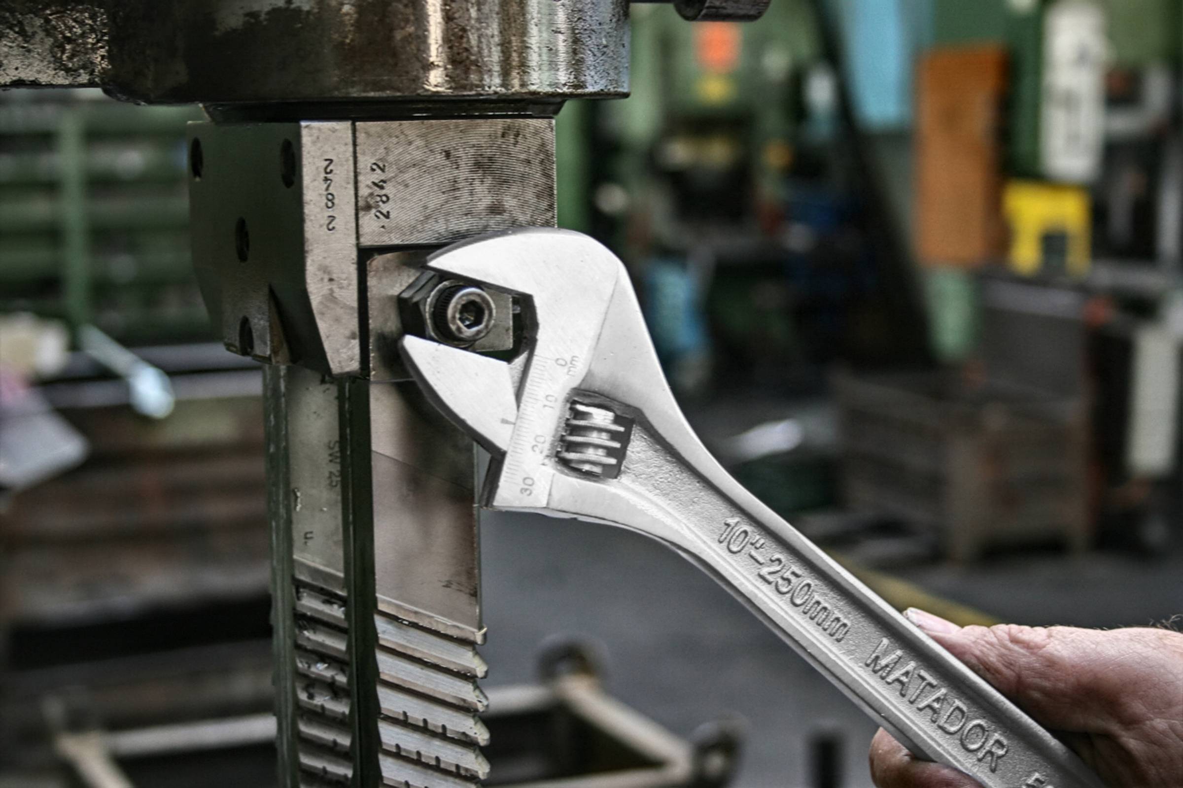 Nastawny klucz płaski odkręca śrubę w metalowej maszynie w warsztacie. Otoczenie ma charakter przemysłowy.