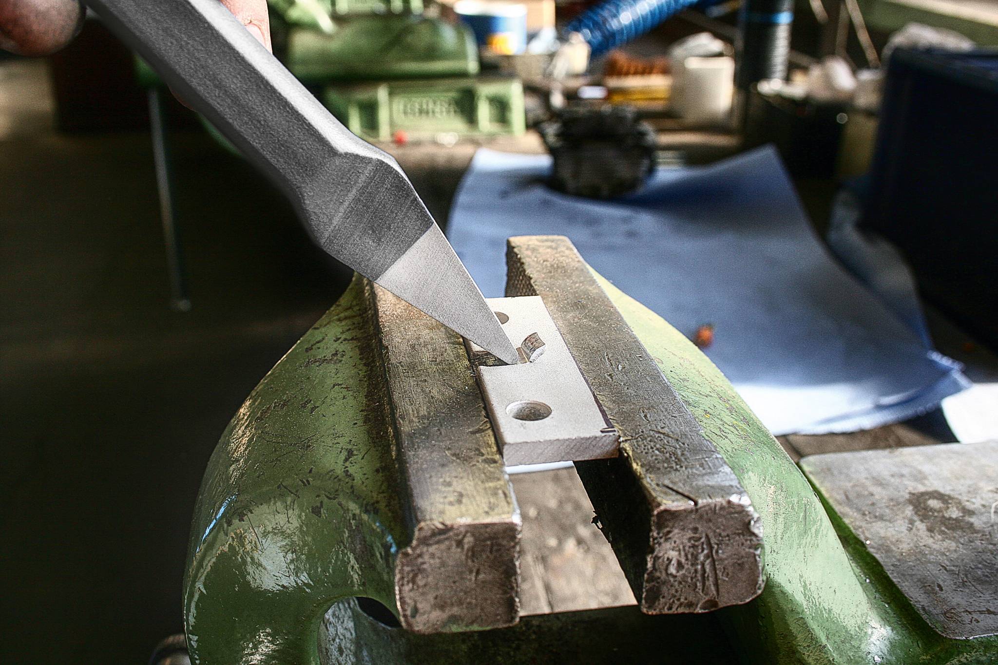 Dłuto obrabia kawałek metalu, który jest zaciśnięty w imadle na warsztacie. W tle widoczne są kolejne narzędzia.