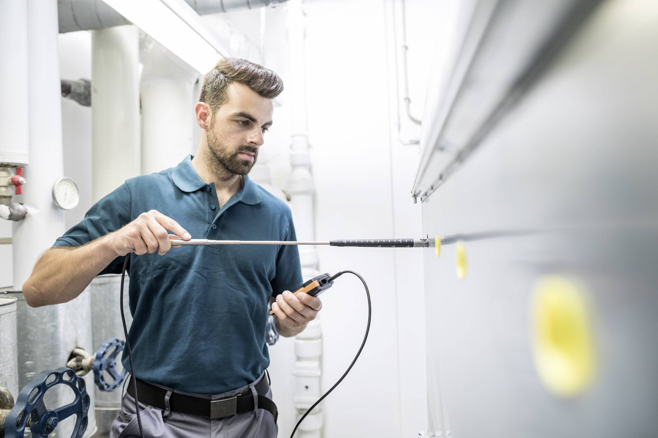 Mężczyzna w pomieszczeniu technicznym mierzy temperaturę ściany za pomocą elektronicznego urządzenia pomiarowego. Ma na sobie ciemnozieloną koszulę.