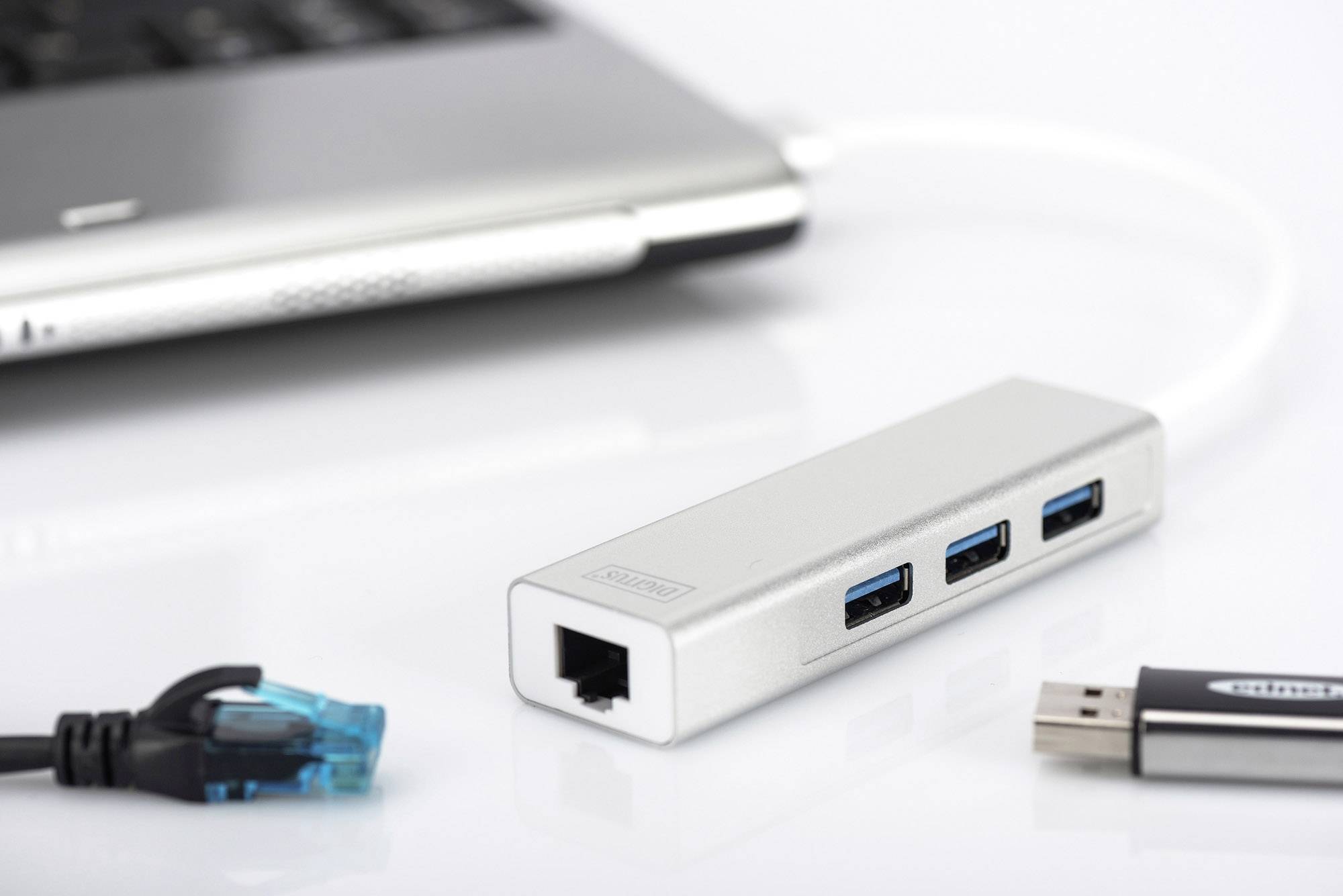 Srebrny hub USB z trzema portami USB i złączem Ethernet leży obok laptopa. Obok niego umieszczono pendrive'a i kabel Ethernet.