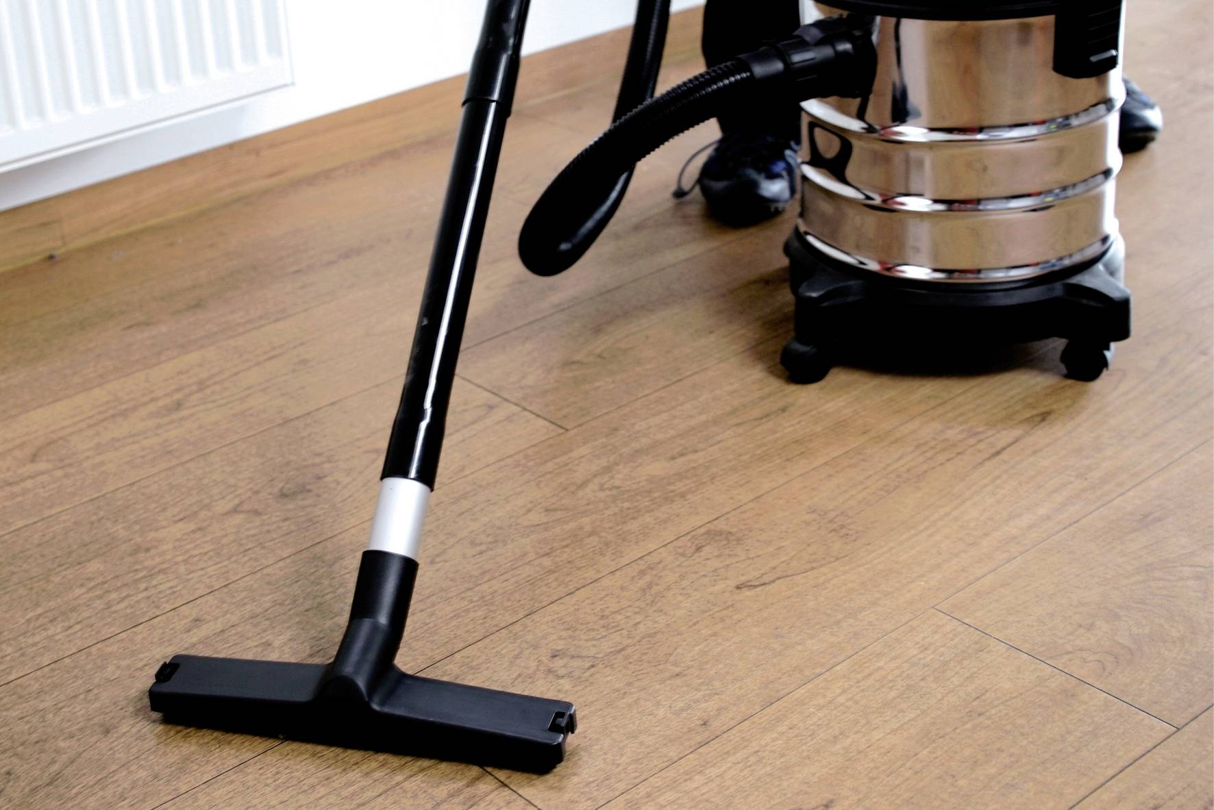 Osoba czyści drewnianą podłogę czarnym odkurzaczem. Pomieszczenie wygląda czysto i schludnie.