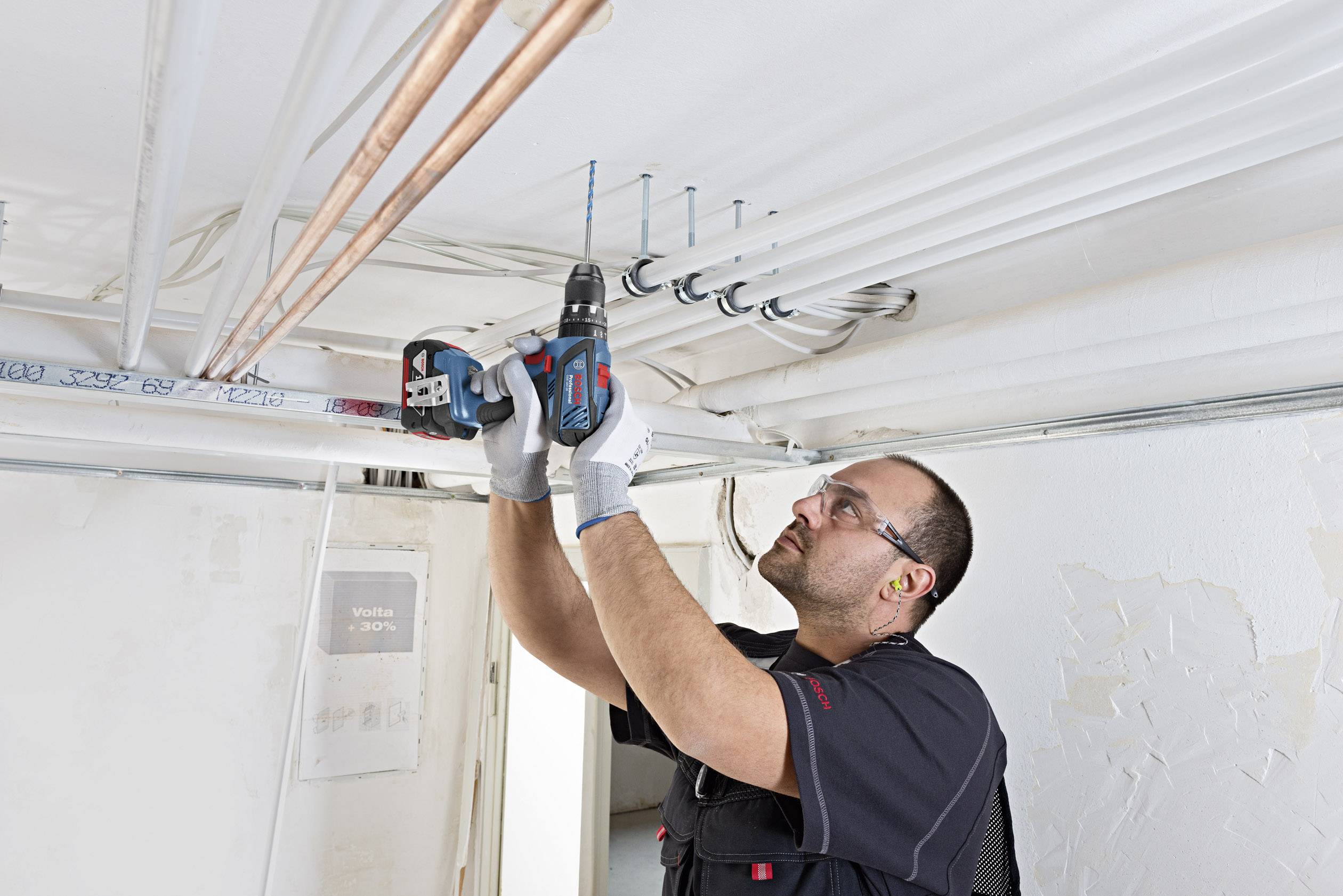 Rzemieślnik wierci w suficie za pomocą wiertarki, aby zamocować rury. Ma na sobie okulary ochronne i rękawice.