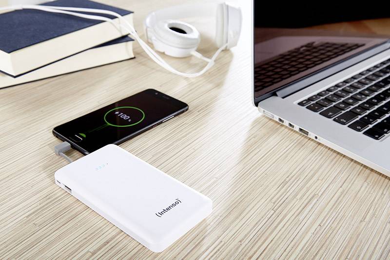 Biały powerbank ładuje smartfon wyświetlający 100% poziomu baterii. Obok leży laptop i słuchawki.