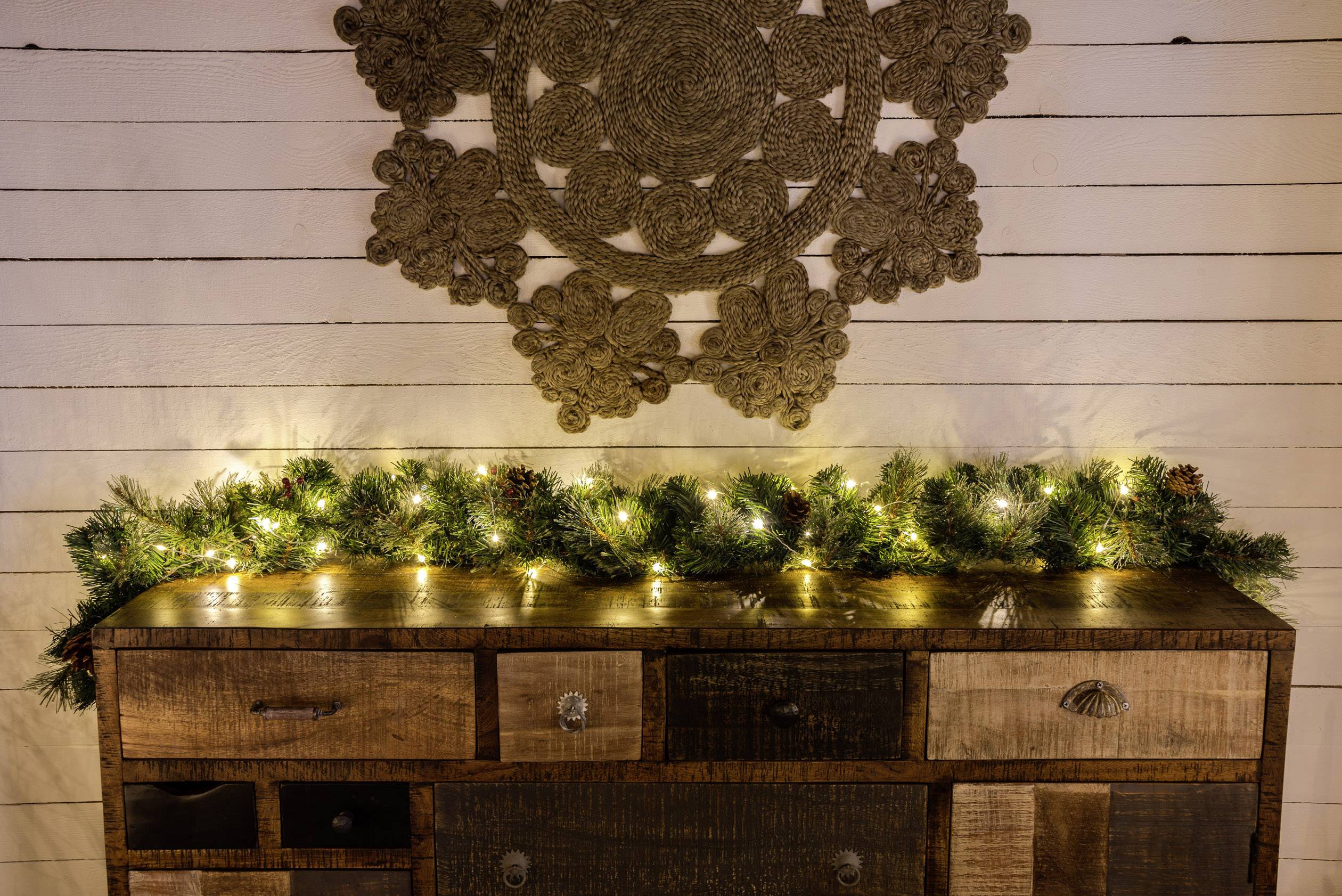 Rustykalna komoda przed białą drewnianą boazerią, ozdobiona girlandą świerkową i ciepłymi lampkami.