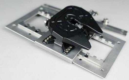 Czarny, mechaniczny zawias zamocowany na metalowej płycie. Wygląda na urządzenie służące do mocowania lub regulacji.