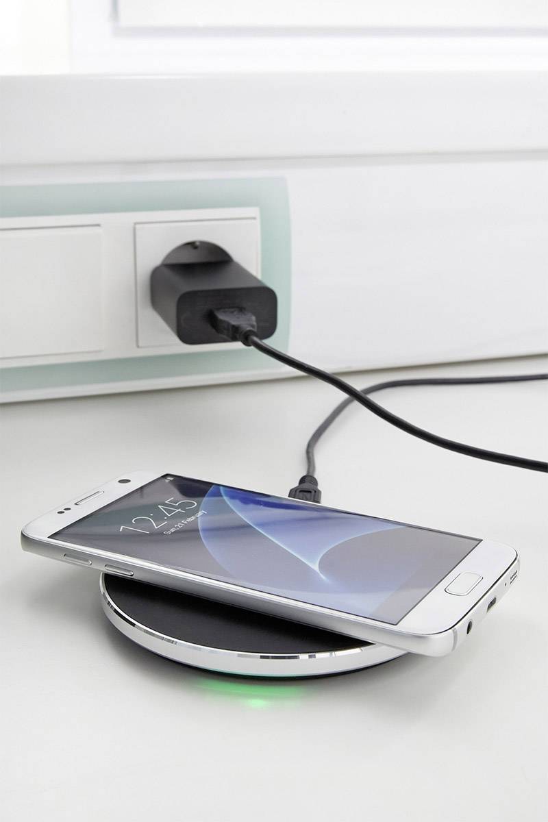 Smartfon leży na bezprzewodowej ładowarce, która jest podłączona przewodem do gniazdka elektrycznego. Na ekranie widać godzinę '12:45'.