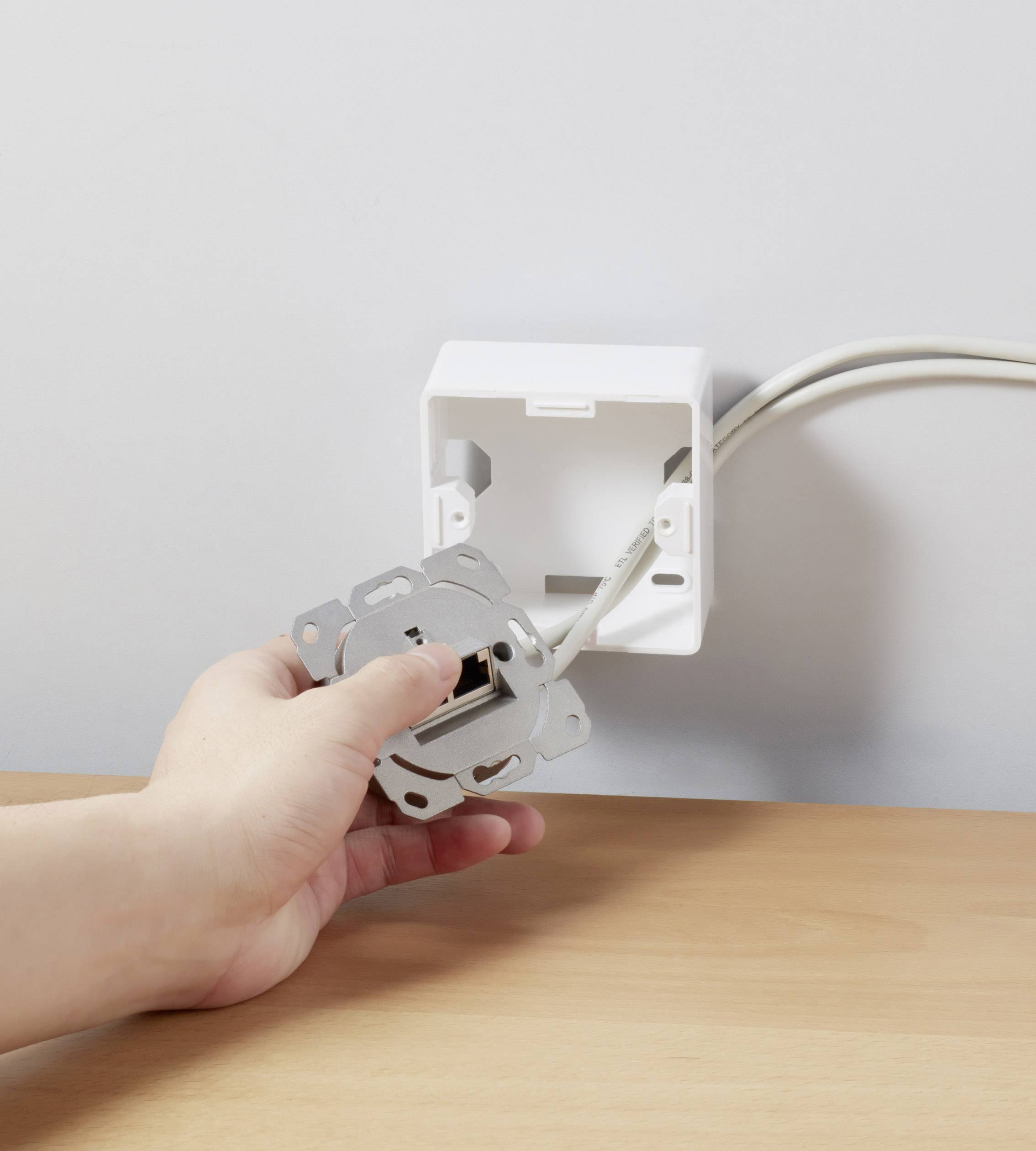 Ręka instaluje gniazdko elektryczne w puszce podtynkowej. Z ściany wystają dwa przewody, które są podłączane do gniazdka.