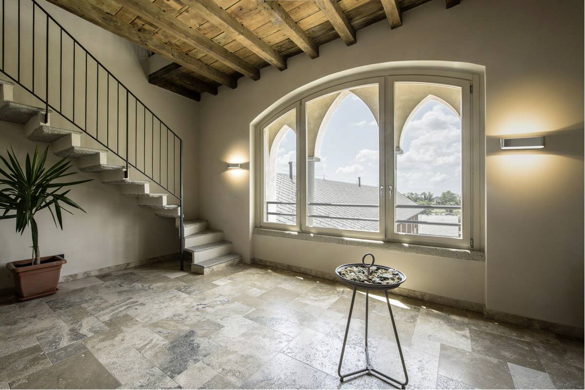 Przestronny pokój zalany światłem, z kamienną podłogą, rośliną w rogu, schodami po lewej stronie oraz okrągłym stołem ustawionym przed dużym oknem w kształcie łuku.