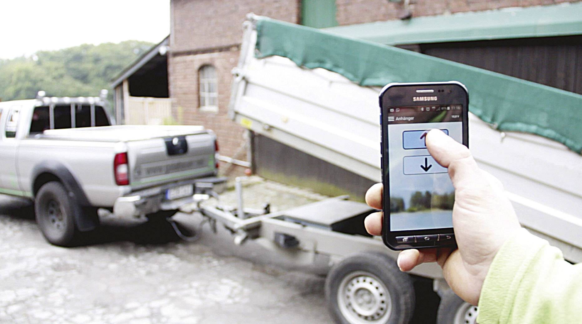 Osoba steruje hydrauliczną funkcją przechyłu przyczepy za pomocą smartfona. Na tle widoczny budynek i pojazd.